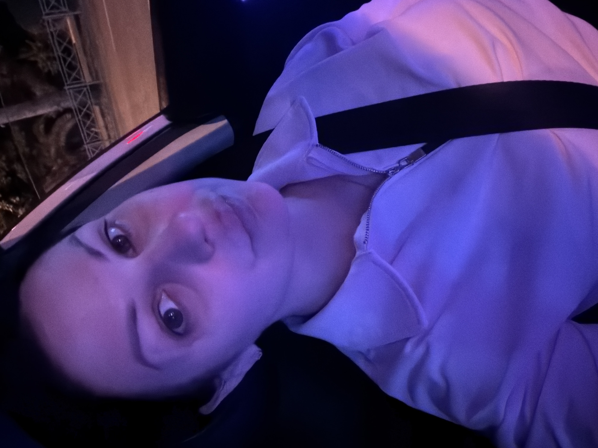 A young woman with dark hair and dark eyes is sitting in the passenger seat of a car at night. She is wearing a white collared shirt with a zipper and a black seatbelt is fastened across her chest. The car interior is dimly lit, with purple and pink lighting casting shadows on her face and clothing. Outside the car window, a street scene is visible, with streetlights, trees, and a fence. The location is identified as Sochi, Russia.