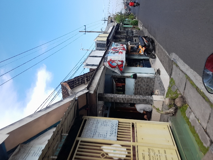 The scene is a street in Kecamatan Tanggulangin, Indonesia, with coordinates -7.505 latitude and 112.718 longitude. There are several buildings on the street, including a shop with a sign that says "99". The street is paved and there are power lines overhead. There is a motorcycle parked on the street. There is a man sitting on the ground in front of a building. There is vegetation growing on the ground and on the side of the street. The sky is blue with some clouds.