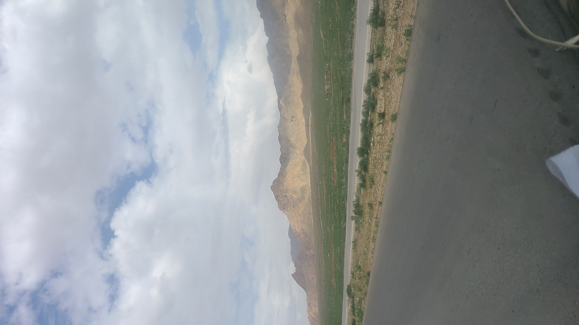 The image displays a vast outdoor landscape under a partially cloudy sky. The sky, occupying the left side of the frame, is filled with large, white and light grey clouds, with patches of clear blue sky visible in between.
In the mid-ground, a long, rugged range of barren mountains stretches across the horizon. These mountains are predominantly light brown and tan, showcasing a dry, rocky terrain devoid of significant vegetation, characteristic of an arid or semi-arid region.
The foreground is dominated by a dark grey, paved road or highway that runs diagonally from the bottom right towards the center. Adjacent to the left side of this road is a prominent strip of vibrant green land, which appears to be cultivated fields or lush pastures, contrasting sharply with the parched, sandy earth surrounding it and the distant mountains. Sparse, low-lying green vegetation is scattered along the edges of the road and on the dry ground. On the far right edge, a small white object, possibly a piece of cloth or paper, and some thin, dark lines, possibly cables, are partially visible. The perspective suggests the photograph was taken from a vehicle.
The scene depicts a wide-open, natural environment in a mountainous area. The lighting indicates it is daytime, likely late morning or afternoon, given the diffused light filtering through the clouds. No people or specific activities are visible. The landscape is consistent with the geographical characteristics of Khanai Baba, Pakistan. No visible text is present in the image.
