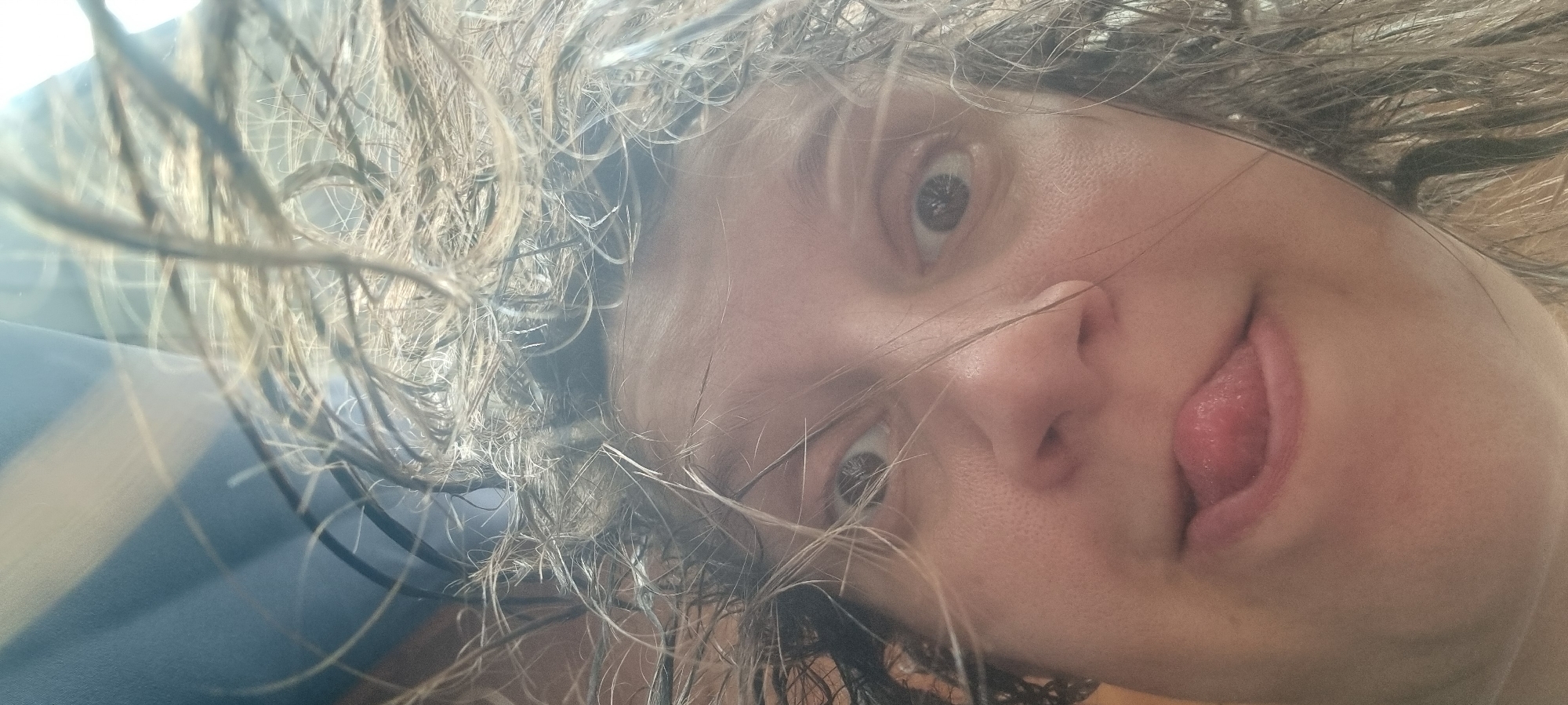 A close-up, slightly blurry selfie of a person's face, tilted diagonally. The person has wide eyes and is sticking their tongue out. Wet hair is framing the face, obscuring some features. The background is indistinct with a hint of blue. Location information is not discernible from the image.