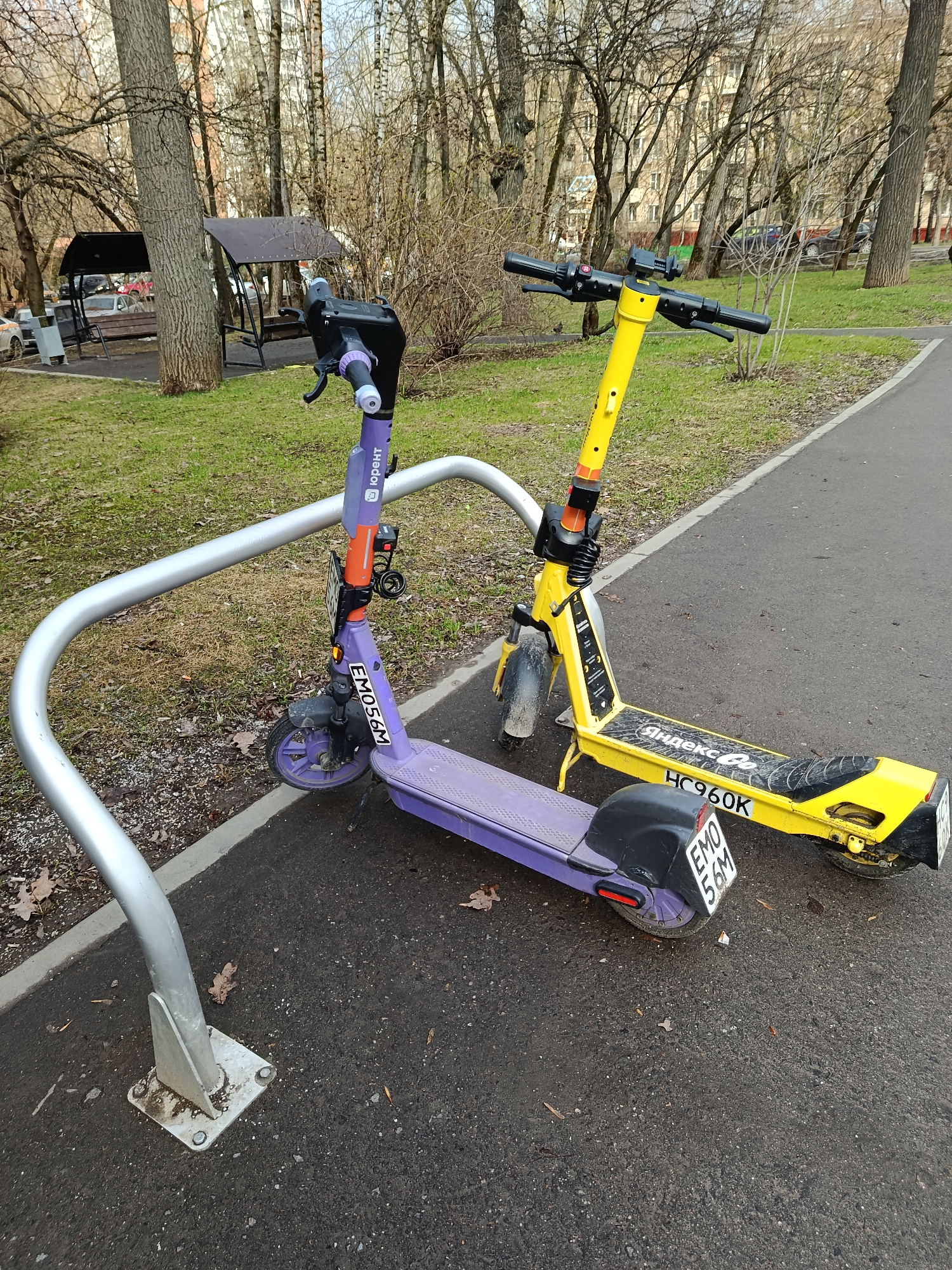 The image shows two parked electric scooters in a park in Moscow, Russia. The scooters are positioned beside a curved metal bike rack.
One scooter is yellow and has the "Яндекс Go" logo on its side, with the license plate "HC960K". The other scooter is purple with the license plate "EMO56M".
The background includes a paved path, grass, bare trees, a gazebo, and a building in the distance. The overall scene suggests an early spring or late autumn day in a park setting.
