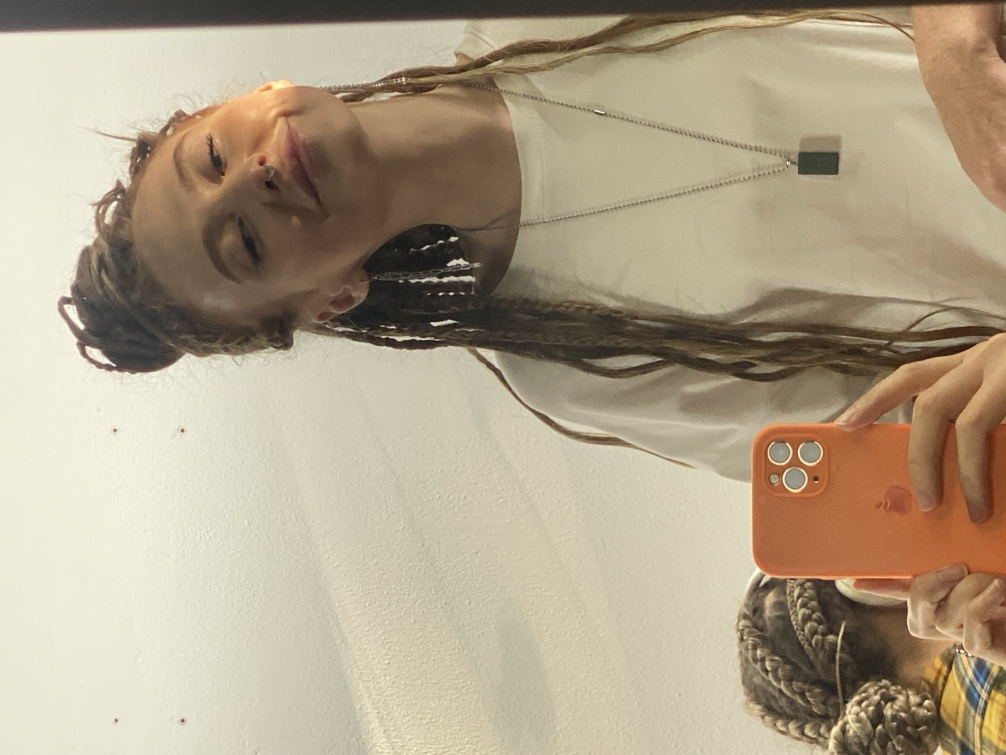 This image captures a mirror selfie of a person in an indoor setting, likely in Sochi, Russia. The primary subject is shown from the chest up, with their head tilted back and eyes softly closed, exhibiting a serene expression with a subtle smile. They have long, light brown hair styled in numerous thick braids or dreadlocks, some of which are swept over their shoulders. A small, metallic nose piercing is visible.
The person is wearing a plain white t-shirt and a silver-colored chain necklace with a rectangular, dark green or black pendant. Their left arm is extended upwards, holding an orange-cased smartphone, likely an iPhone given the visible three-camera lens array on the back and the faint outline of the Apple logo on the case. The phone is pointed towards the mirror, capturing the reflection.
In the bottom right corner, a partial reflection of another person or part of the primary subject's lower body is visible, showing more braided hair styled in a bun and a yellow plaid shirt. The background consists of a plain, light-colored wall, possibly white or off-white, with some minor surface imperfections and small dark spots. The lighting appears soft and even, suggesting indoor artificial light or an overcast day. No specific time of day or weather conditions can be determined. The overall atmosphere conveyed is calm and contemplative.