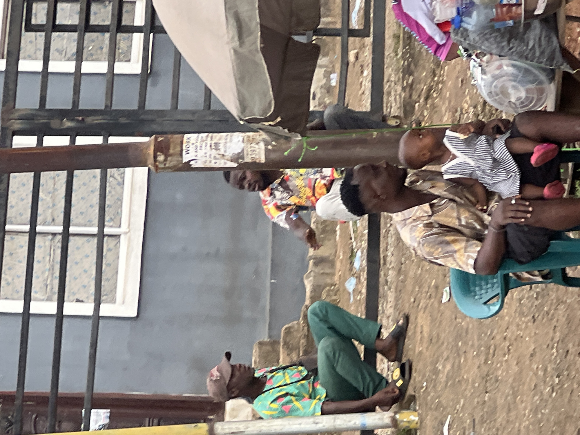 The image depicts a street scene in Nkpor, Nigeria, with several individuals in the foreground and background. In the lower center, a man wearing a patterned shirt and black shorts is holding a young child dressed in a striped dress and pink shoes. They are seated, with the man looking to his right. To the left, a man wearing a green patterned shirt and green pants is seated on steps, looking to his right. He is wearing a baseball cap and sandals. In the middle ground, another man in a brightly colored shirt is standing. A lamppost with posters on it stands between these groups. In the background, a building with windows is visible, and a market stall with an umbrella is partially visible on the right. The ground appears to be dirt and debris. The overall impression is of a bustling urban environment.