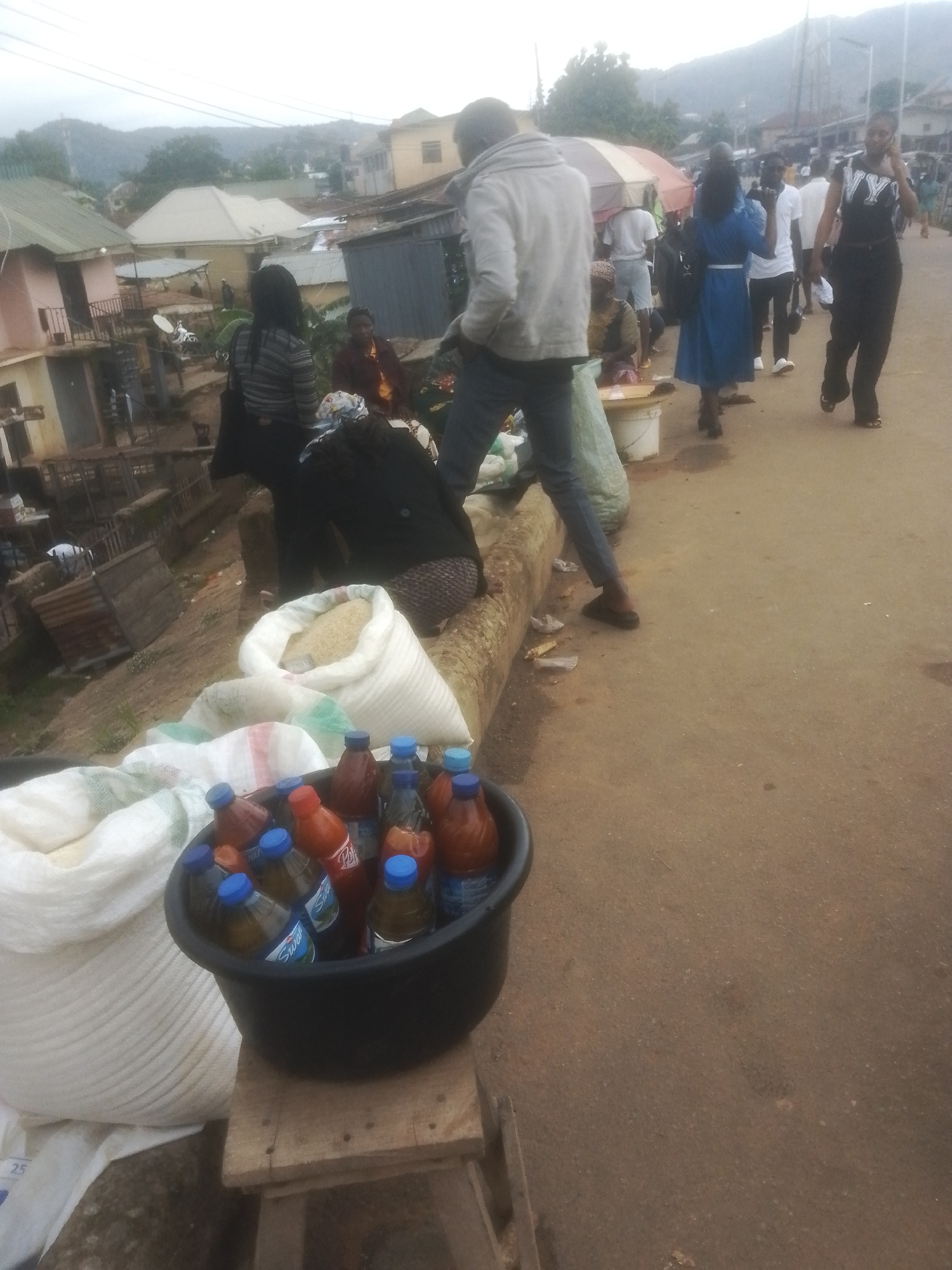 This image depicts a street scene in Gwong, Nigeria, likely during daytime given the ambient light. The setting appears to be a bustling market or roadside area, with a variety of people engaged in daily activities.
In the foreground, a black plastic tub overflows with numerous bottles of what appear to be beverages, many with blue caps. Next to the tub are large white sacks, one filled with what looks like rice, and another partially visible sack with colored stripes. The bottles and sacks are placed on a wooden structure that serves as a stand or display.
Several individuals are visible in the mid-ground and background. A person in a grey hooded sweatshirt and dark pants stands prominently on the left, appearing to observe the scene. Further back, other people are walking and standing, some under a colorful umbrella, suggesting a marketplace. One person in a blue dress is walking away from the camera, while another in a black top with "NY" on it is seen on the right, talking on a phone. A person in a dark hijab and patterned skirt is seated in the mid-ground. The overall atmosphere seems busy and communal.
In the background, buildings with various roof styles and colors line the street, and hills can be seen in the distance, indicating a semi-urban or semi-rural environment. The ground is paved with asphalt. The weather appears to be overcast, contributing to the soft lighting. No specific event is clearly indicated, but the gathering of people and goods suggests a typical market day. No discernible text is clearly visible on the bottles beyond brand names that are difficult to read.