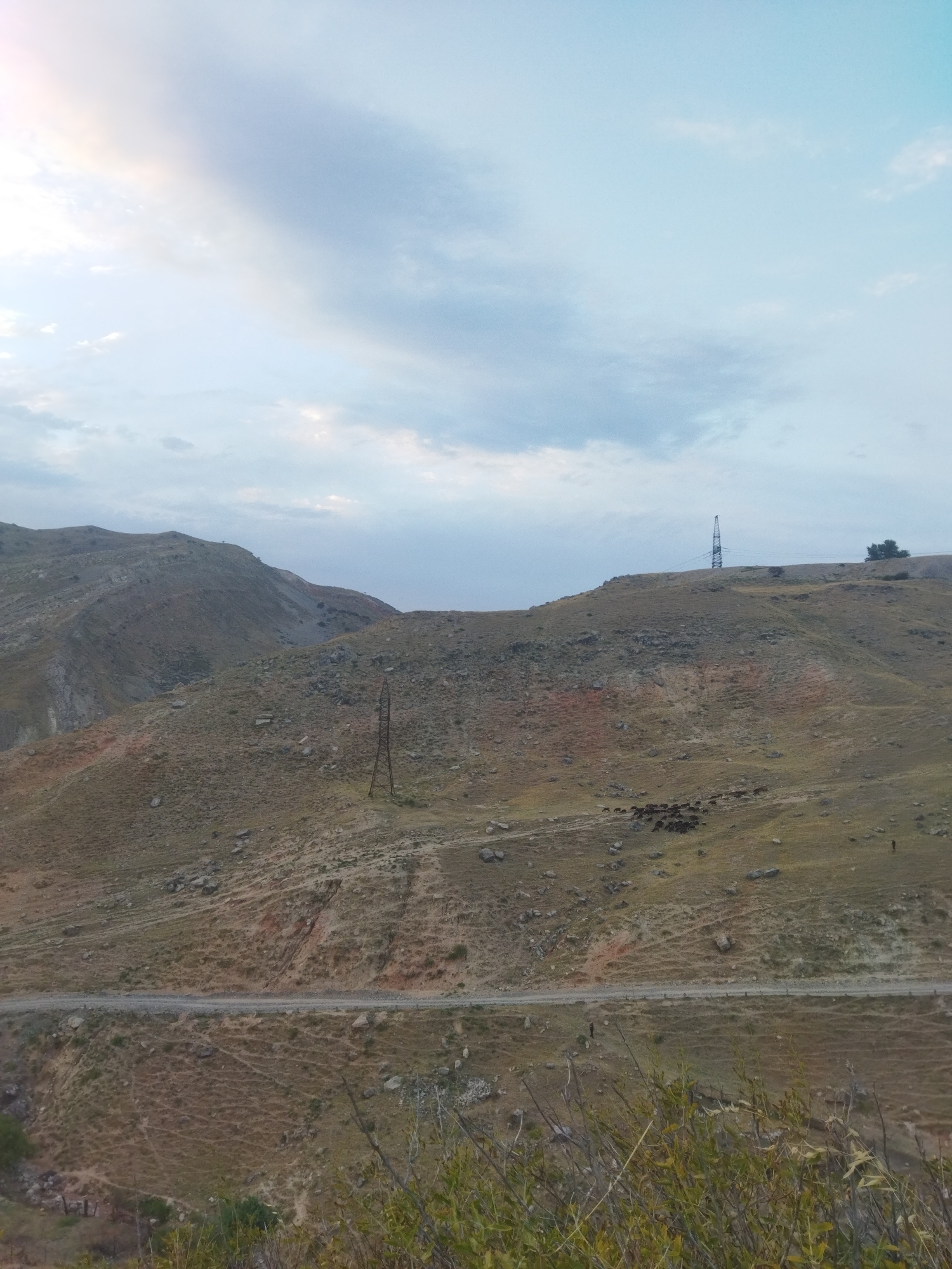 The image depicts a mountainous landscape in the city of Mailuu-Suu, Kyrgyzstan, under a cloudy sky. The terrain is characterized by rolling hills covered in dry grass and scattered rocks. A dirt road traverses the middle ground of the image, with a steep incline on the far side. On the crest of one of the hills, a tall, lattice-like electricity pylon stands prominently, with power lines extending from it. In the distance, a small group of dark-colored animals, possibly livestock, are visible grazing on the hillside. The foreground features some bushy vegetation. The overall impression is one of a rural, somewhat rugged environment.