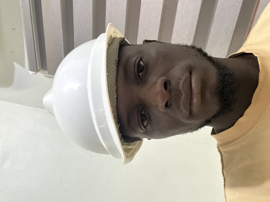 An adult male, with dark skin, a short beard, and mustache, is depicted from the chest up, looking directly forward. He wears a white hard hat, positioned on his head with a visible, light-colored, perforated inner lining showing beneath the brim. He is dressed in a light brown, long-sleeved shirt. The background, an indoor setting within the city of Lafia, Nigeria, features a plain white wall on the left side of the frame and vertical window blinds with alternating light and dark grey horizontal stripes on the right. A white pull cord for the blinds is visible.