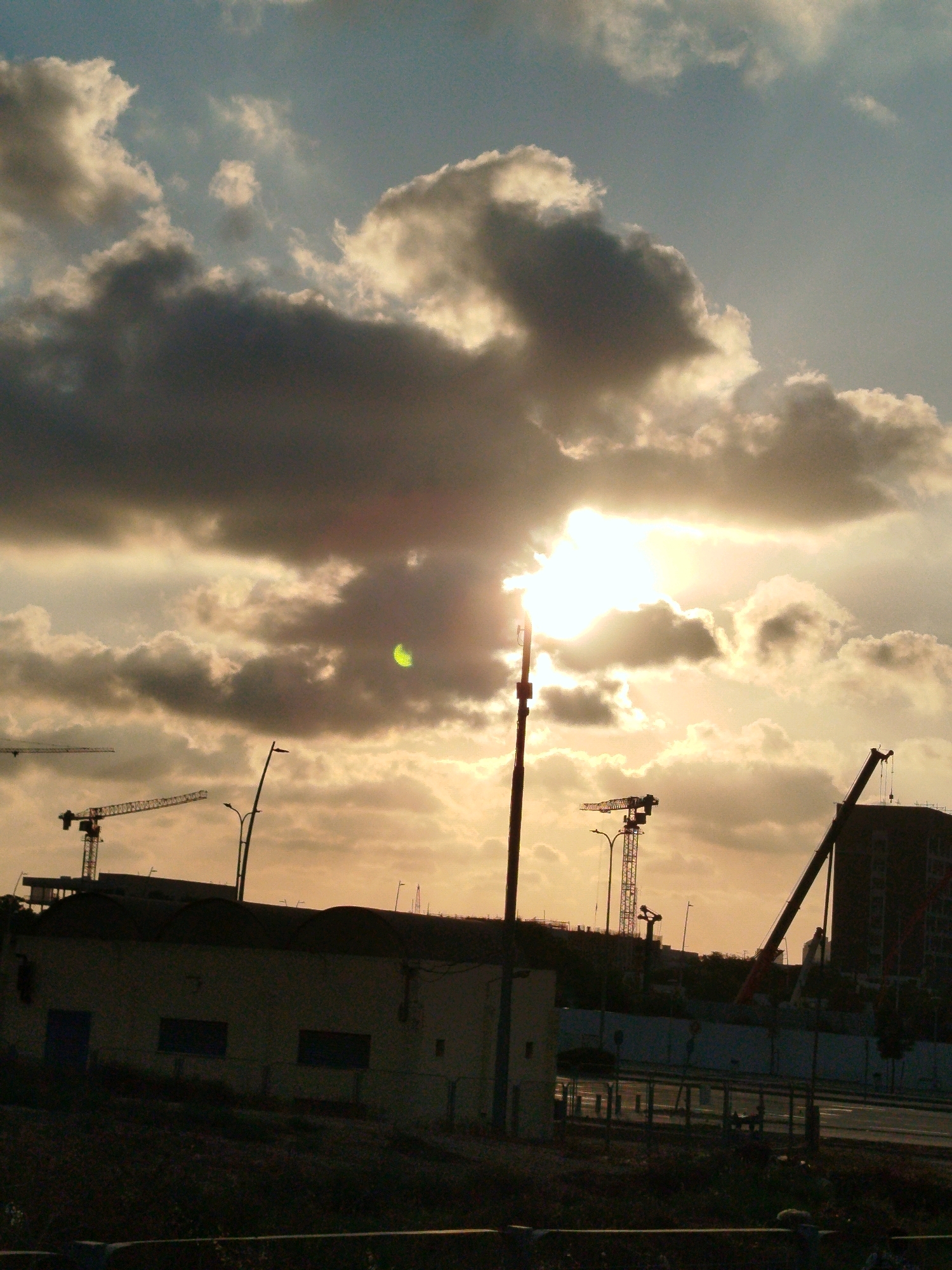 This is a vertical, medium-shot, outdoor photograph taken in Ashdod, Israel, featuring a dramatic sky illuminated by the setting sun. The sky is filled with a mix of dark, ominous clouds and lighter, illuminated clouds, creating a stark contrast. The sun is partially obscured by the clouds, casting a bright glow and lens flare across the upper center of the image.
In the foreground, a dark silhouette of a low-lying building is visible, with arched rooftops. To the left of this building, a construction crane is partially silhouetted against the sky. Further to the right, the dark silhouette of a taller building is visible, with a large red crane extending diagonally upwards. Another smaller crane, a tower crane, is visible between the low building and the taller building. A street light pole is prominently positioned in the center of the image, silhouetted against the sky. The ground in the immediate foreground is dark and indistinct, appearing to be overgrown with vegetation. A fence-like structure runs horizontally across the bottom of the image. The scene appears to be a construction site at dusk.