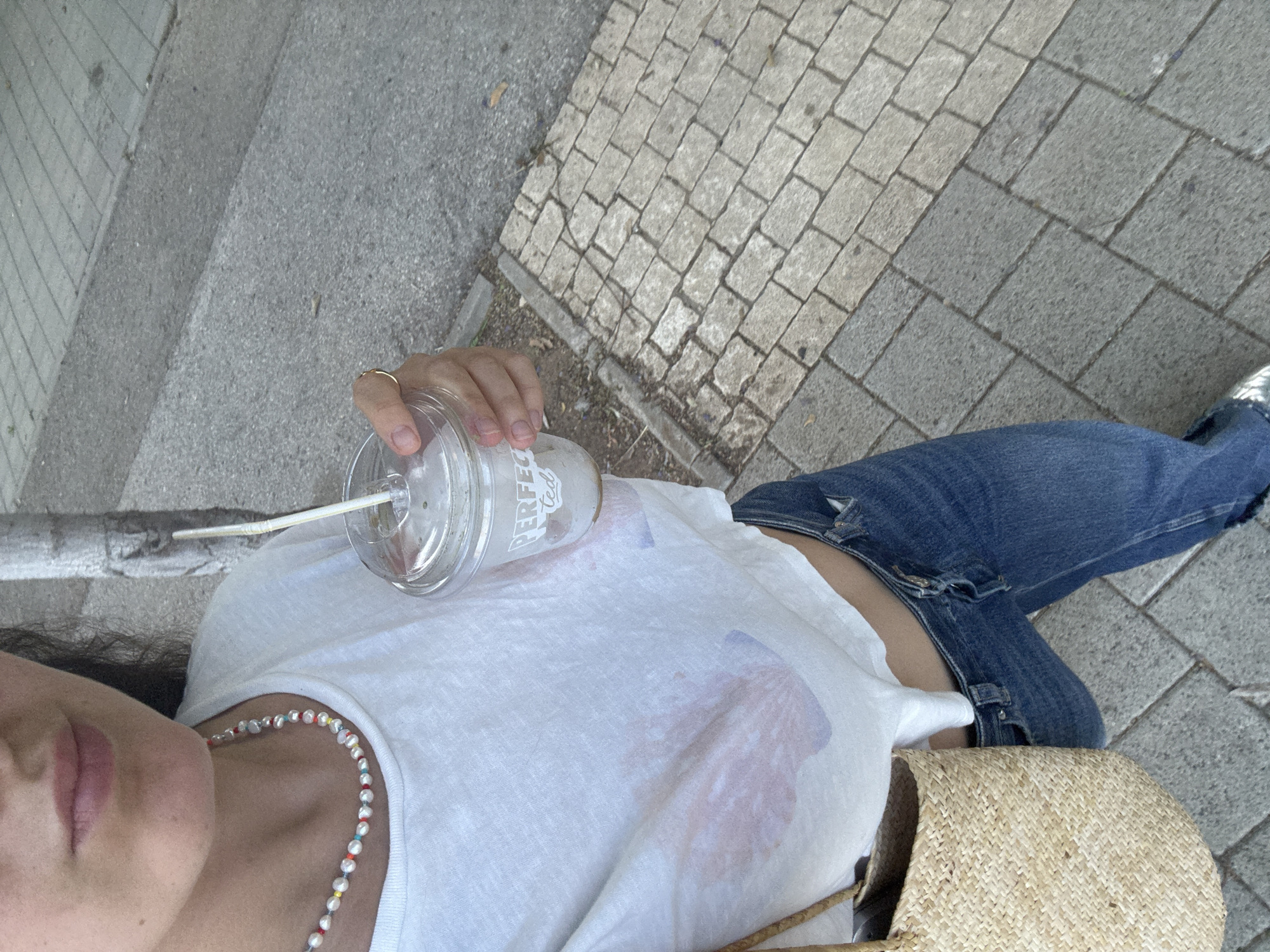 A person stands outdoors on a paved sidewalk. They are holding a clear plastic cup with a white lid and a straw, containing a beverage. The person is wearing a white cropped t-shirt, blue jeans, and a pearl necklace with colorful beads. A woven straw bag is visible on their left side. The background shows a mix of paved surfaces and what appears to be a tree trunk. The scene suggests a casual outing in a city environment, possibly in Herzliya, Israel, given the architectural style of the paving stones and the general ambiance.