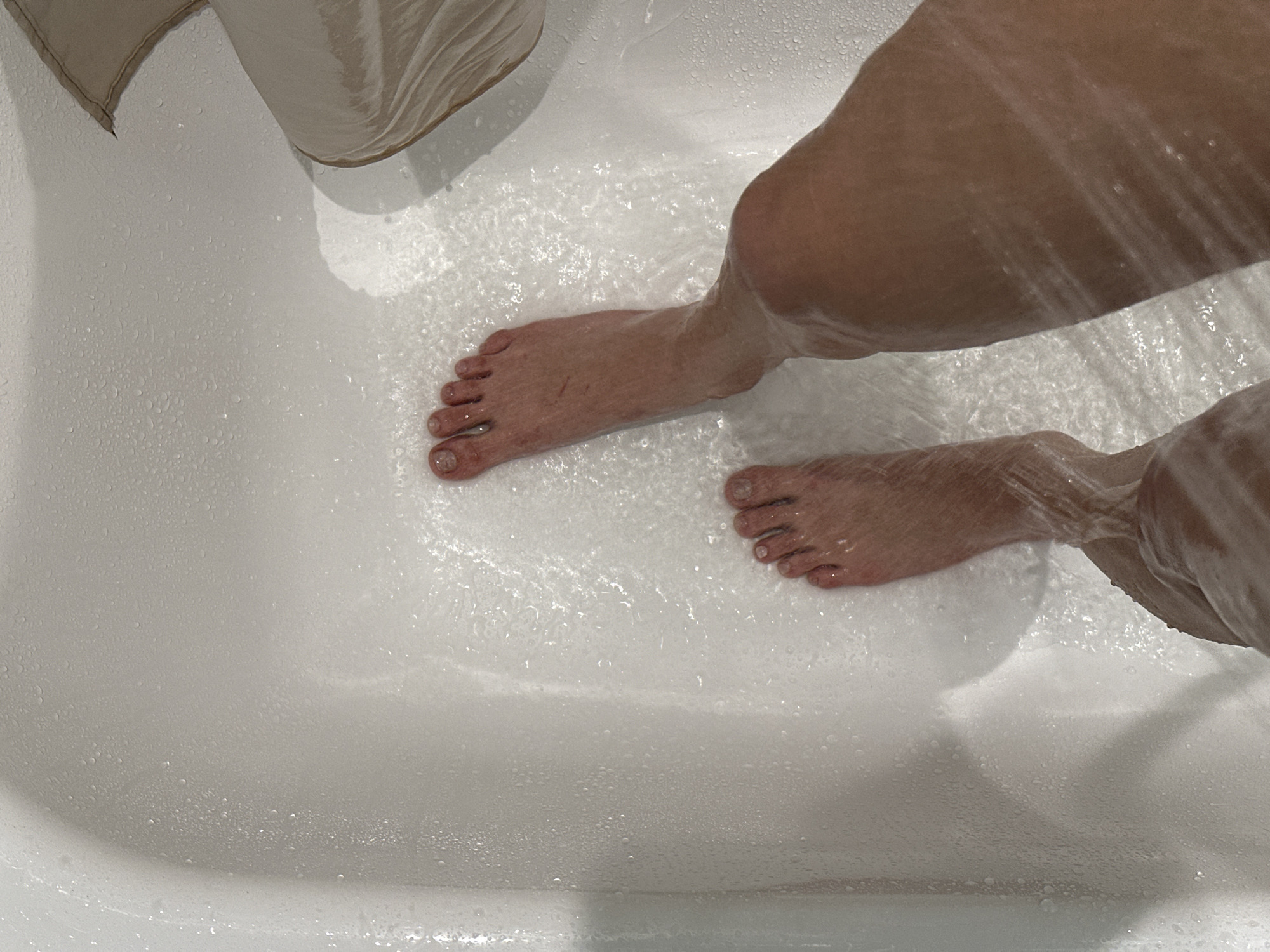 A view from above shows two human feet in a white bathtub under running water. The tub also contains a tan object, potentially a curtain. The context suggests a private residence. There is no information to place the image within the city of Krasnoyarsk, Russia.