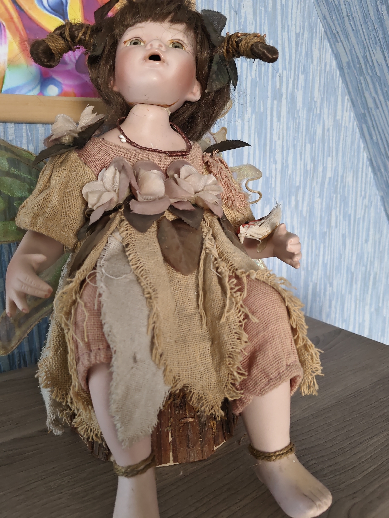 This is a close-up, eye-level, medium shot of a porcelain doll with brown hair styled in two braided pigtails, sitting on a wooden surface. The doll is dressed in a tattered, earth-toned outfit made of burlap and pink fabric, adorned with artificial flowers and leaves. Its neck appears broken, and its mouth is open. The doll's arms and legs are visible. In the background, a blue, textured wall and a colorful abstract artwork are present. The scene is likely an indoor setting. No specific location in Rostov-on-Don, Russia can be identified from the image.