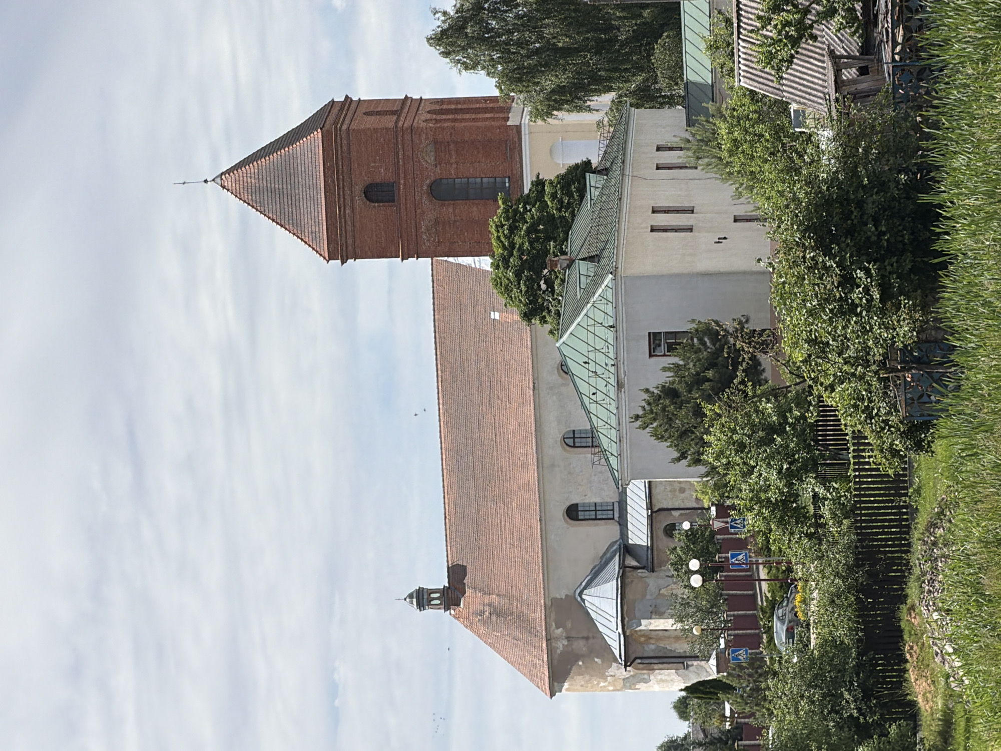 The image depicts a vertical shot of a historical building complex, likely a church with an adjacent tower. The architecture is primarily brick with terracotta tiled roofs and some lighter colored sections. The tower is tall and rectangular, made of red brick with arched windows. The main building has a long, sloping tiled roof and lighter colored walls with visible windows. There are sections of green metallic roofing visible, suggesting modern renovations or additions. Surrounding the buildings are lush green trees and vegetation. A dark wooden fence runs along the right side of the image, partially covered in ivy. In the background, a bright, slightly overcast sky with wispy clouds is visible. Several street signs with blue backgrounds and white symbols are positioned along a path or road near the buildings. There is a parked car visible behind the fence. The scene appears to be a daytime setting, with good natural light. No people are directly visible in the image. The overall impression is one of historical architecture set within a well-maintained, green environment.