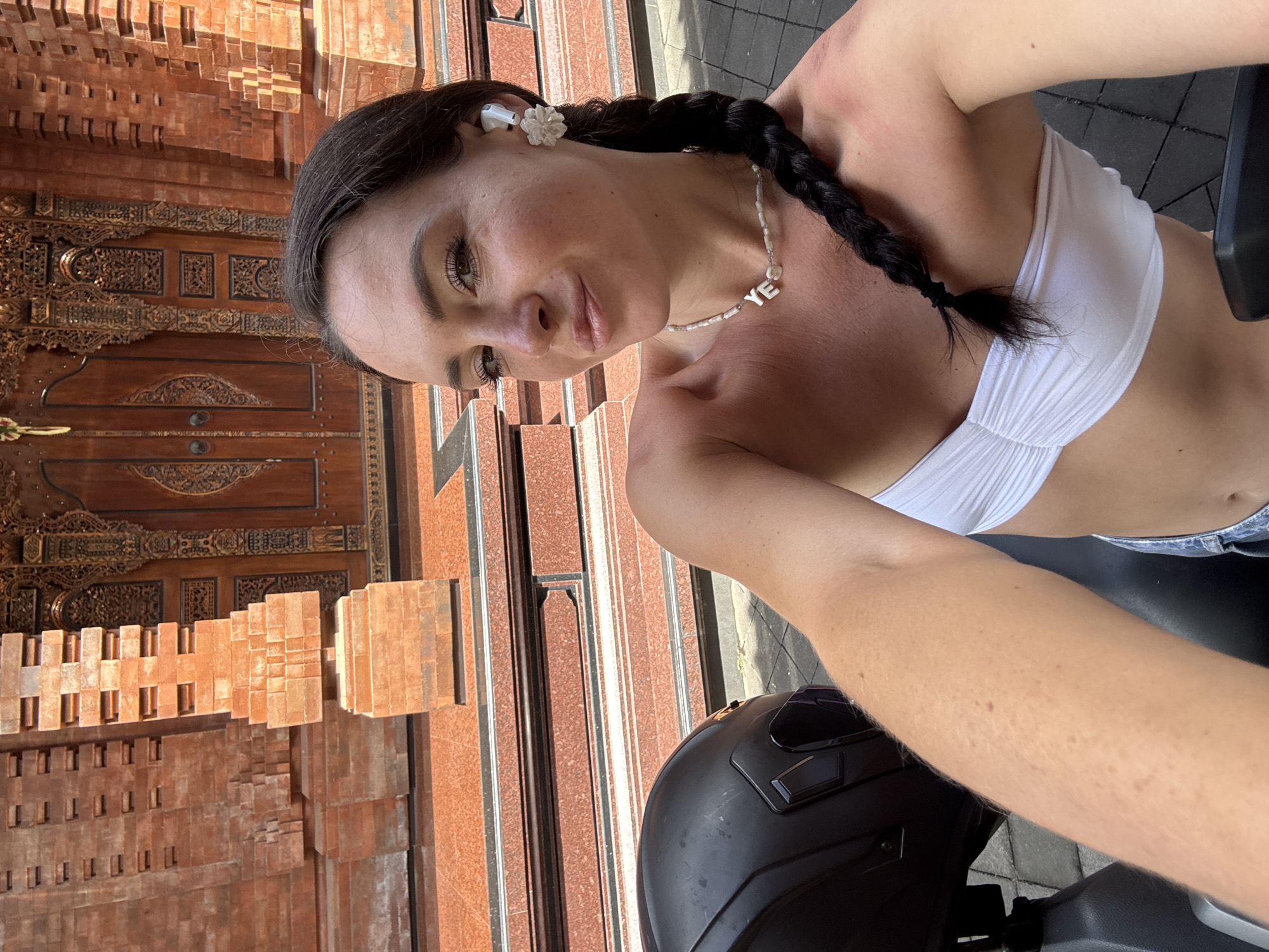 In North Kuta, Indonesia, a woman with dark hair braided to her side and wearing a white bandeau top takes a selfie outdoors. She is positioned in front of a large, ornate wooden door flanked by red brick structures. A black motorcycle helmet is visible to her left. She is wearing a pearl necklace with the letters "YE" and a white flower-shaped earring in her right ear. Sunlight illuminates the scene, casting shadows and highlighting the textures of the brickwork and the woman's skin.