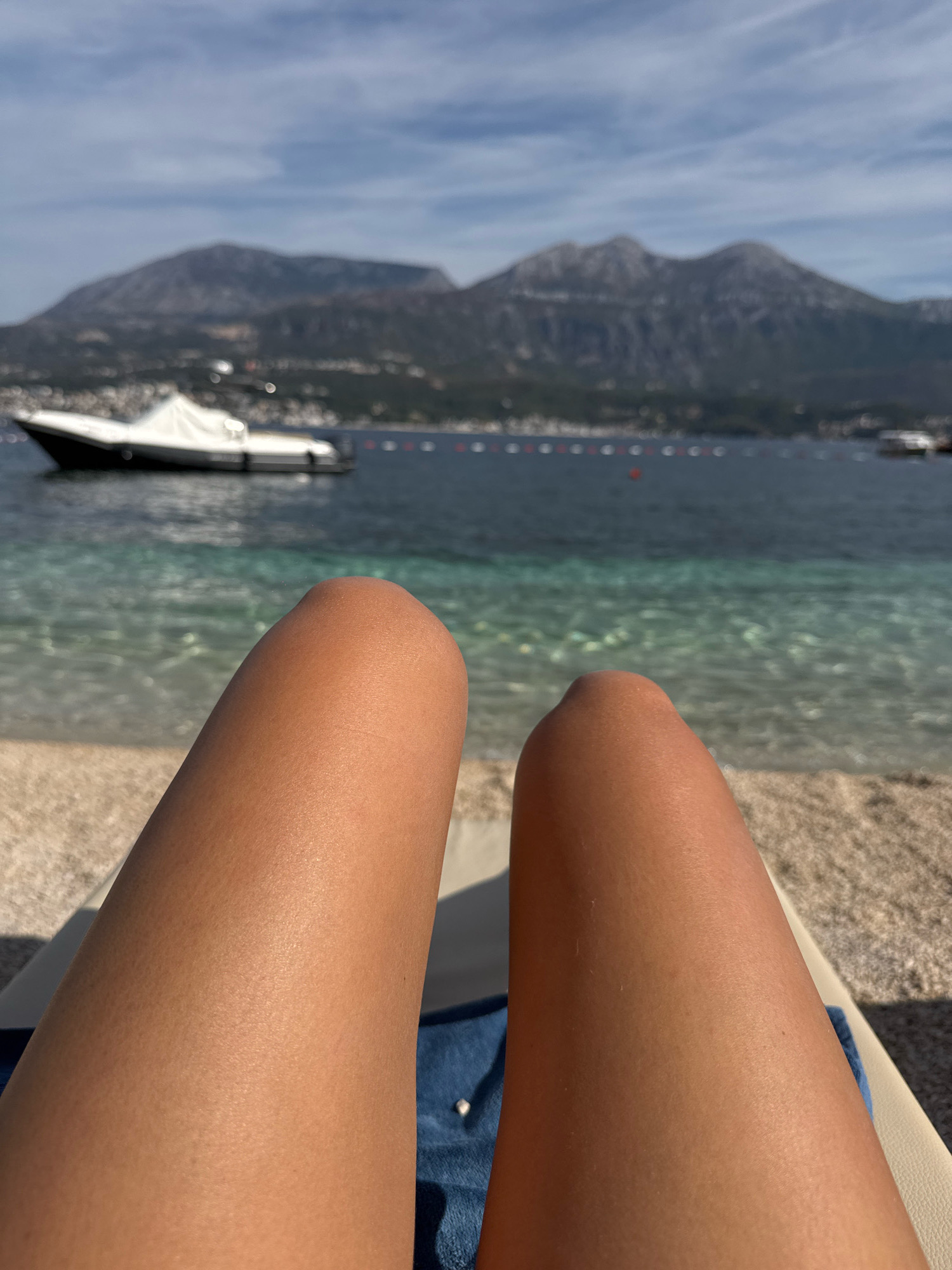 A first-person perspective shot of a person's tanned legs on a beach chair shows a beautiful scenic view of the sea and mountains. The clear turquoise water laps gently onto the pebble beach, and in the distance, a sleek white boat is anchored. The majestic mountains rise in the background, their peaks softened by the hazy sky. The sun-drenched scene evokes a sense of relaxation and vacation.
