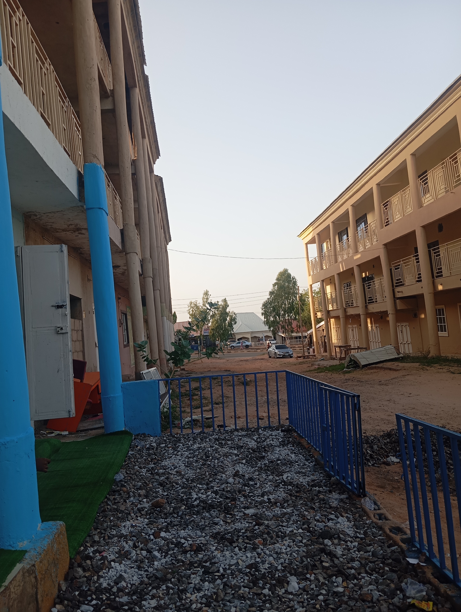 The image depicts an outdoor scene in Gombe, Nigeria, located between two multi-story buildings. The building on the left has exposed concrete columns painted partially in blue. The building on the right has beige walls and white railings on the balconies.
In the foreground, there is a fence, painted blue, with an area covered in grey gravel. Beyond the fence, the ground appears sandy and unpaved. In the background, vehicles and distant buildings are visible. The sky is clear and light in color, indicating either early morning or late afternoon lighting. No people are visible in the frame.