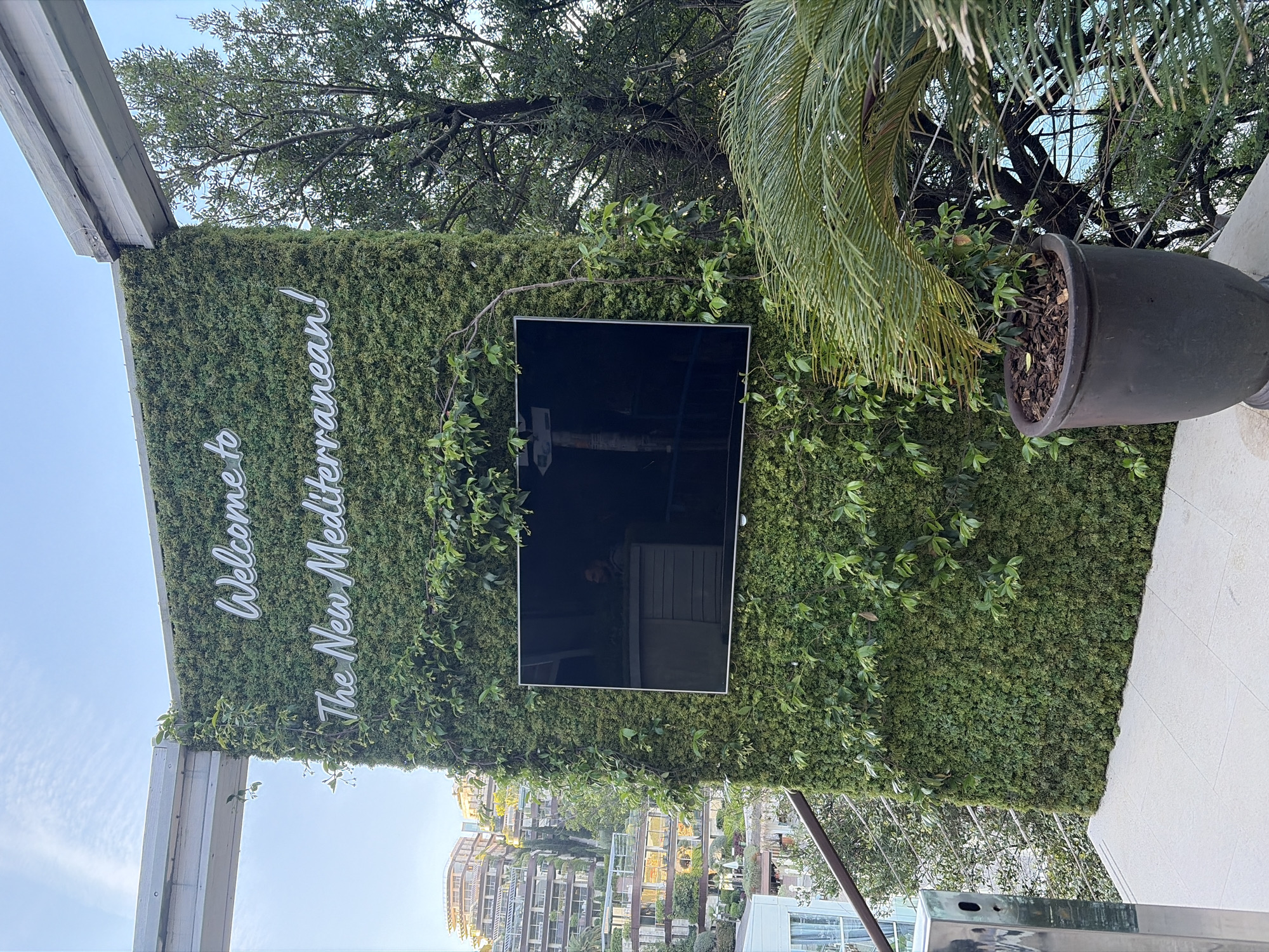 Here is a description of the image:
The image shows a vertical garden or green wall, possibly in an outdoor setting. The sign is in English, reading "Welcome to the New Mediterranean!" There is a flat-screen television mounted on the green wall. The surrounding area includes trees, plants, and what appears to be a building complex. Based on the visual cues, it is likely located in Budva, Montenegro, as the architecture and vegetation match typical scenes from the region. The overall impression is that of a modern, possibly upscale, outdoor venue, such as a restaurant or event space.