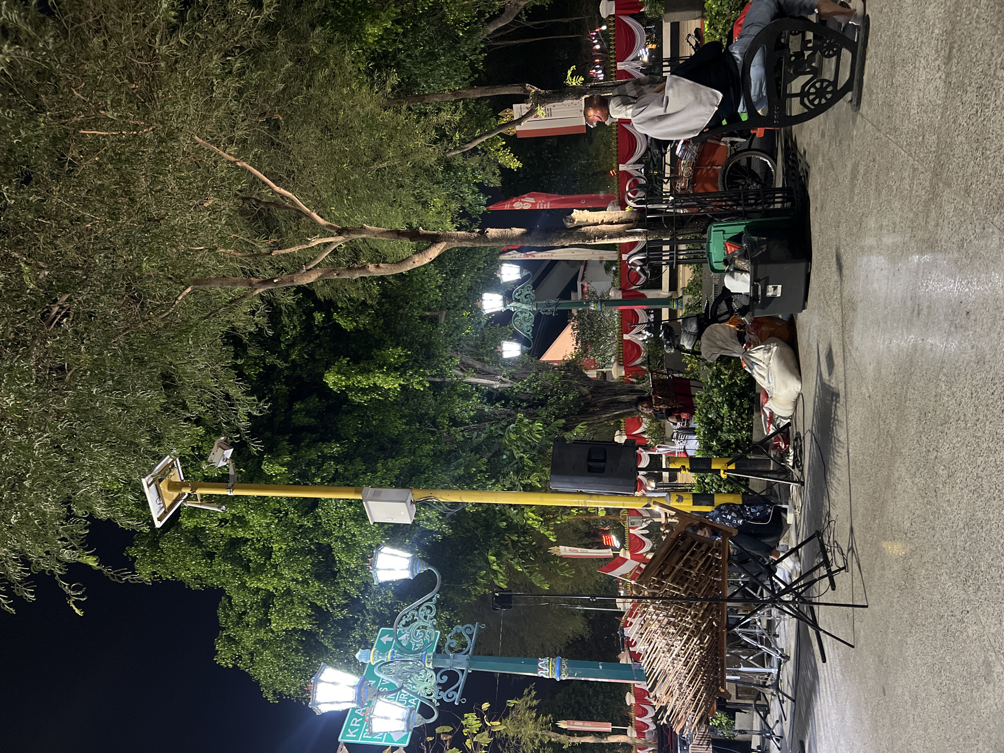 The image depicts an outdoor night scene in Malioboro Street, Yogyakarta, Indonesia. A performance setup is visible, featuring a large wooden stringed instrument (likely a gambus or similar lute) on a stand in the foreground, surrounded by musical equipment including speakers and cables. In the background, a street lamp with multiple ornate lights illuminates the area, with a solar-powered surveillance camera mounted on a yellow pole. Red and white drapes are strung along a railing, suggesting a festive or formal occasion. Several people are present, some seated and others potentially involved in the performance or observing. The overall atmosphere appears to be lively, with the warm glow of the lights contrasting with the dark night sky.