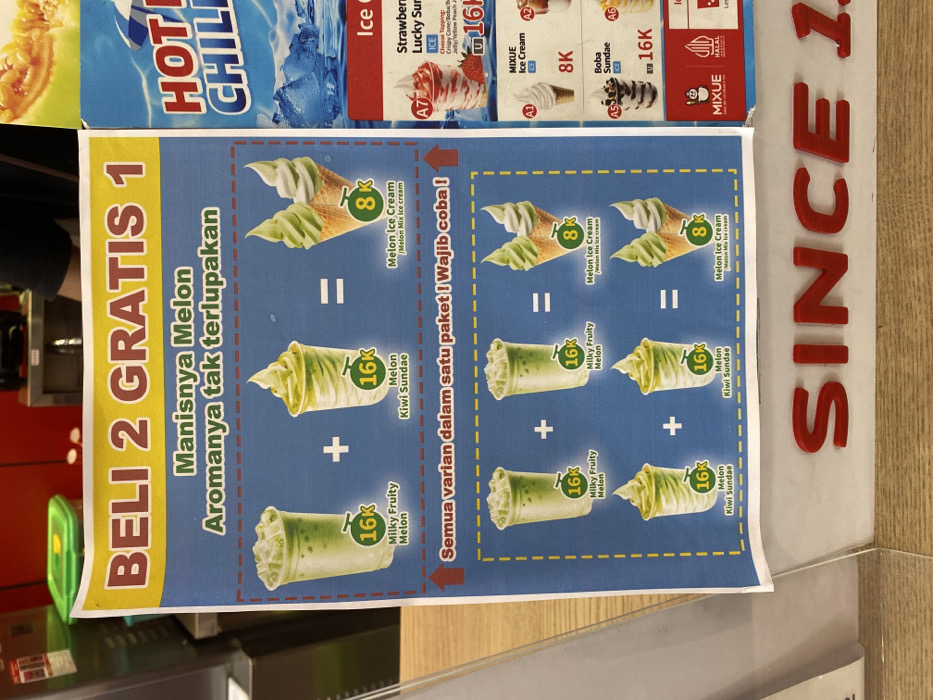 The image shows a promotional poster for a "Beli 2 Gratis 1" (Buy 2 Get 1 Free) offer for melon-based desserts. The poster is displayed in a shop located in Kecamatan Piyungan, Indonesia, at the coordinates -7.837 latitude and 110.474 longitude. The poster features three different dessert combinations, each consisting of two 16k (Indonesian Rupiah) drinks and one 8k melon ice cream. The drinks are labeled as "Milky Fruity Melon" and "Melon Kiwi Sundae." The ice cream is labeled as "Melon Ice Cream / Melon Mix Ice Cream." The poster also mentions "Semua varian dalam satu paket! Wajib coba!" (All variations in one package! Must try!). The poster is displayed in a shop with a wooden counter, and is likely intended to attract customers to purchase these desserts.