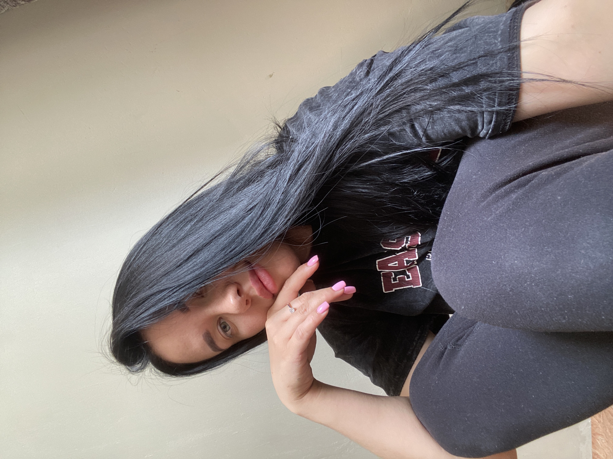 A young woman with long, dark hair and light-colored eyes is sitting indoors. She is wearing a black t-shirt and dark leggings. Her left hand is resting on her cheek, and she has a ring on her left ring finger and brightly colored pink fingernails. Her gaze is directed downwards and slightly to the left. The background appears to be a plain, neutral-colored wall. The image was taken in the city of Chaikovsky, Russia.