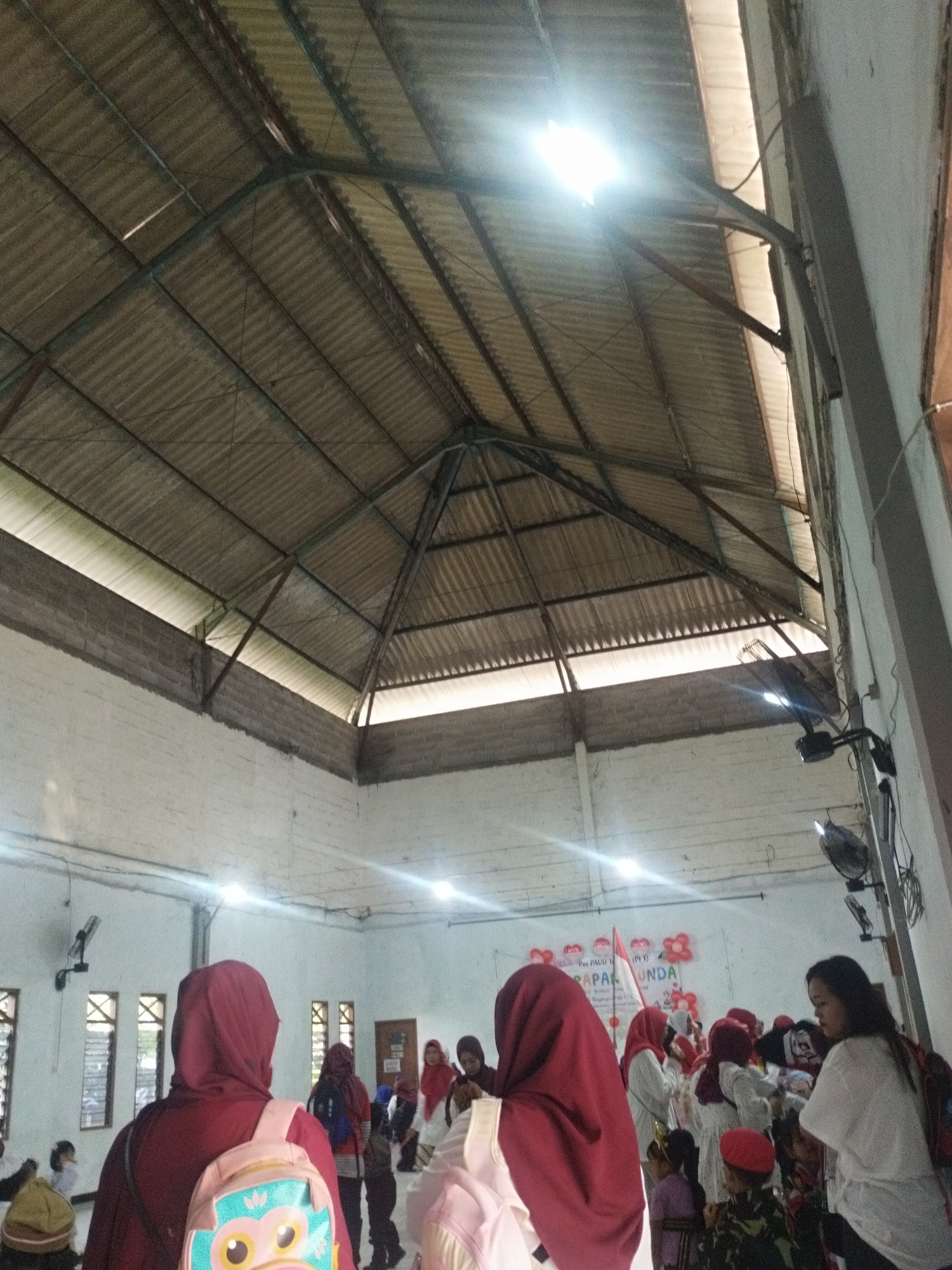 The image depicts an indoor gathering, likely a community event or educational program, taking place in a large, open room with a corrugated metal roof supported by a visible metal frame. The walls are white and appear to be constructed of brick or concrete. Several windows with criss-cross gratings are visible along one wall. In the foreground, a person wearing a maroon hijab and a backpack with an owl design is facing away from the camera, looking towards a group of people in the background.
The background shows a collection of individuals, including children and adults, some of whom are also wearing hijabs. A banner with text that appears to be in Indonesian, possibly reading "POS PAUD TERPADU" and "LUPUNDA" or a similar phrase, is displayed on a wall, adorned with red balloons and an Indonesian flag. The lighting in the room is a mix of natural light filtering through gaps in the roof and artificial lighting from ceiling fixtures and mounted lights. The overall atmosphere suggests a lively event. The scene is consistent with the type of community hall or educational facility found in various neighborhoods across Surabaya, Indonesia, a city known for its vibrant cultural events and community gatherings.