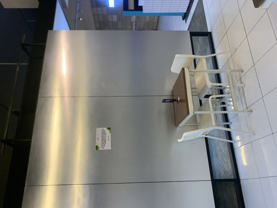 The image shows a small table and two white chairs in a cafe in Malang, Indonesia (coordinates -7.941, 112.608). The table is made of wood and has a brown tabletop. The chairs are made of metal and have white seats. The cafe has white tiled floors and a wall made of stainless steel. There is a sign on the wall with text in Indonesian. The sign is not visible enough to decipher what it says. The cafe is relatively empty, with no people present in the scene. The cafe is located in a building with black metal support beams. There is a window in the background that allows a glimpse of the outside world. The scene is lit by overhead lighting. There is also a small black box on the table. The table is centered in the image. The chairs are positioned on either side of the table. The floor is clean and white. The scene is mostly empty, with no people in the frame.