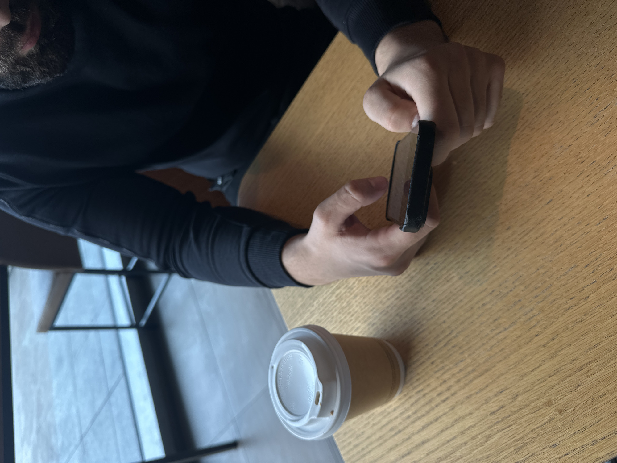 A man with a beard, wearing a black long-sleeved shirt, is holding a black smartphone in his hands, presumably looking at the screen. He is sitting at a light brown wooden table next to a disposable coffee cup with a white lid. In the background, there is a window with a geometric frame. It is impossible to determine the exact location in Sofia, Bulgaria without further clues.
