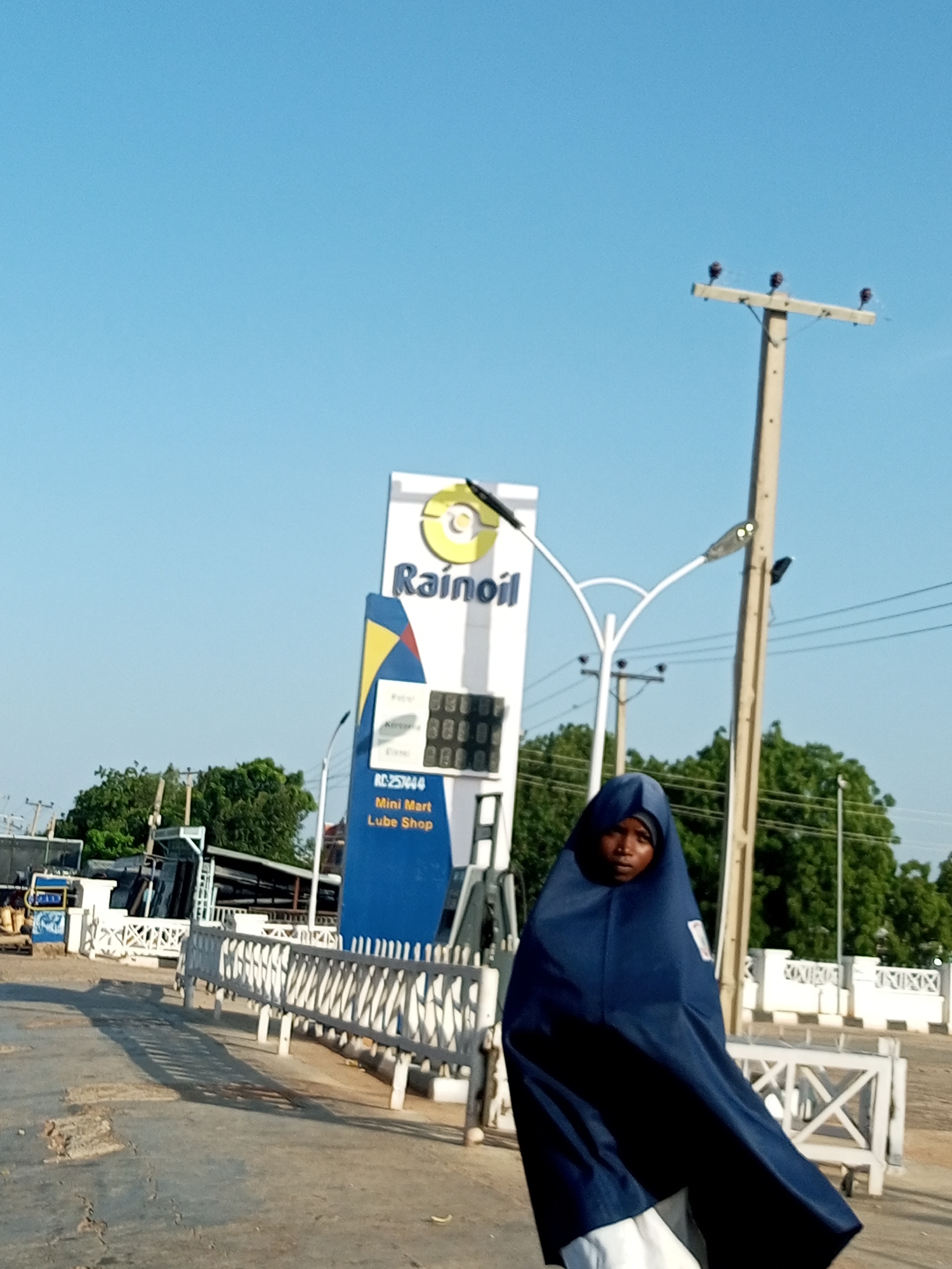 The image depicts a young person wearing a blue hijab and a white undergarment, standing outdoors in what appears to be a public area in Garko, Nigeria. Behind the person is a Rainoil gas station with a prominent white sign featuring the company logo and text indicating "Mini Mart Lube Shop." The gas station has a digital display showing numbers. Several utility poles with insulators are visible, along with trees and a white metal fence. The ground is paved and shows signs of wear. The sky is clear and blue. The scene suggests a typical day at a roadside service station in a Nigerian town.