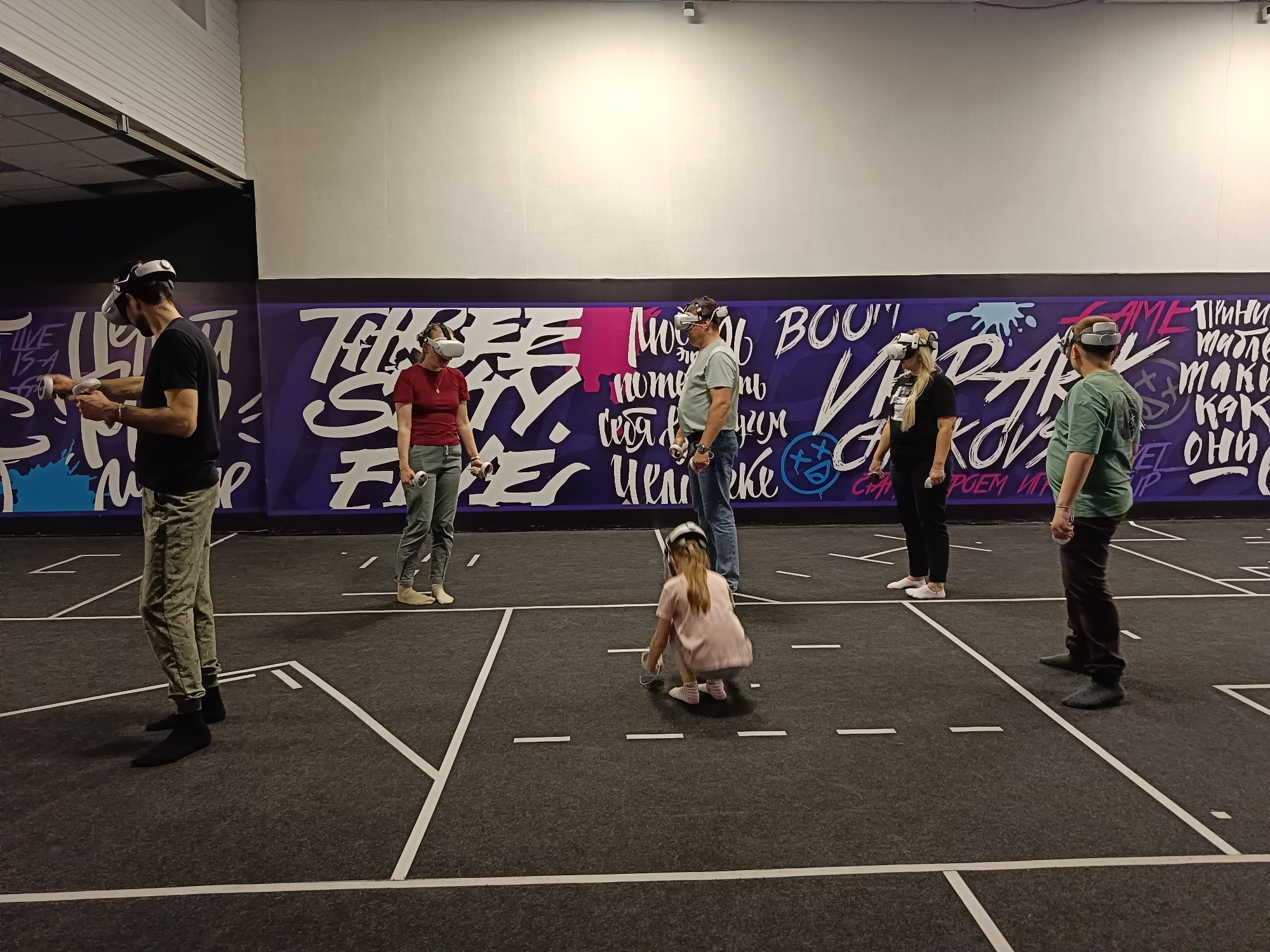 The image depicts an indoor virtual reality (VR) gaming arcade or dedicated play space in Chaikovsky, Russia. Seven individuals are present: six adults and one young child, all actively engaged in a VR experience. Each person is wearing a white VR headset, likely a Meta Quest 2 or similar model, and holding corresponding controllers. The young girl is crouched on the floor, also with a headset on.
The setting features a spacious room with a dark grey carpeted floor marked with white lines, clearly delineating separate play areas for participants. The background is dominated by a vibrant purple wall mural adorned with graffiti-style text in white, pink, and light blue. Above the mural, the wall is plain white, leading up to an industrial-style ceiling with exposed pipes and lighting fixtures.
Visible text on the mural includes English phrases such as "LIVE IS A GAME" and "GAME." Prominent Cyrillic text can also be seen, including phrases like "МОЖЕШЬ ПОТЕРЯТЬ СЕБЯ В ДРУГОМ МИРЕ" (You can lose yourself in another world), "БУМ" (Boom), "ЧТО ТАКОЕ ЧУВСТВА" (What are feelings), and "КАК МЫ ИГРАЕМ" (How we play). The time of day is not discernible due to the indoor, artificially lit environment. The scene portrays an active and immersive entertainment activity.