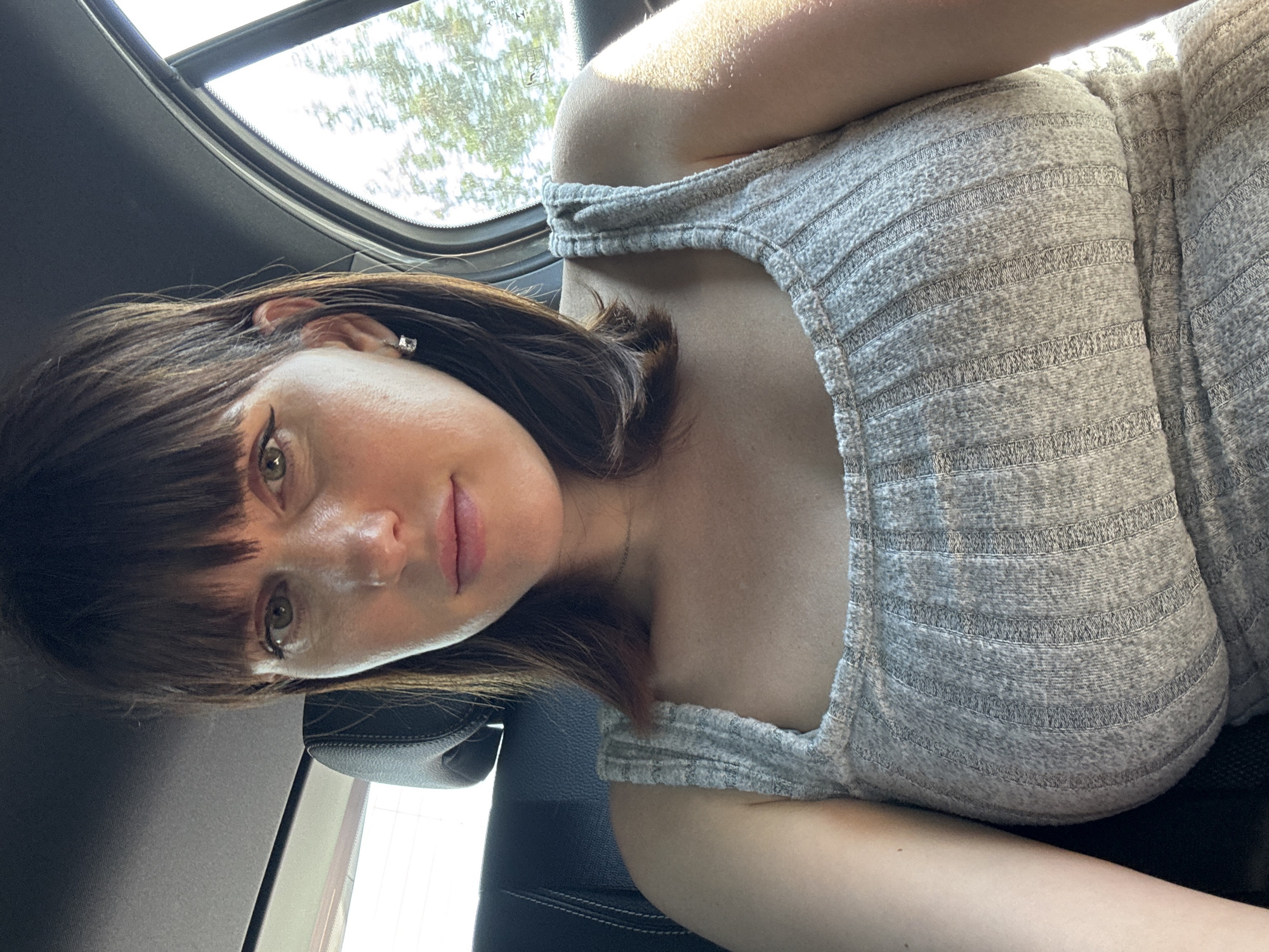 A woman with brown hair and bangs is seated in a car, likely near Pomezia, Italy, though specific landmarks are not visible through the car window. She is wearing a light gray, striped top. The car's interior is visible, including a black leather seat and a sunroof. The lighting suggests it is daytime.