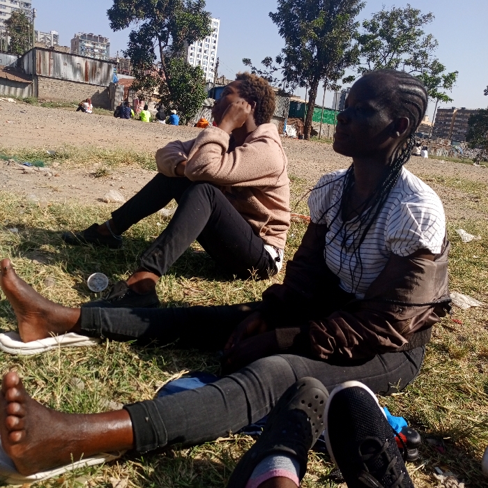 The scene depicts two individuals seated on a sparse dirt and grass patch under a clear, sunny sky, characteristic of an informal settlement bordering a developed urban area in Nairobi, Kenya.
In the foreground, a person with dark skin and braided hair is seated, wearing a white and black striped short-sleeve shirt, a dark jacket draped over their shoulders, and black jeans. Their legs are extended, with the right foot bare and resting on a white sandal sole, and the left foot partially visible, wearing a black athletic shoe with a white sole and a grey sock. This individual's gaze is directed upwards and to the right.
Adjacent and slightly behind, a second person with dark skin and short, curly hair is seated, wearing a light brown hooded sweatshirt and black jeans. This individual's right hand is near their chin, and their gaze is directed upwards and to the left. A small, round, silver-colored object lies on the ground near their left foot.
The mid-ground features a collection of makeshift structures constructed from corrugated metal and concrete, with several other indistinct individuals observed sitting or standing among them. In the distant background, multiple modern high-rise buildings are visible, confirming an urban periphery. The ground throughout the scene is uneven, with patches of dry grass and exposed soil, and appears to have scattered debris.