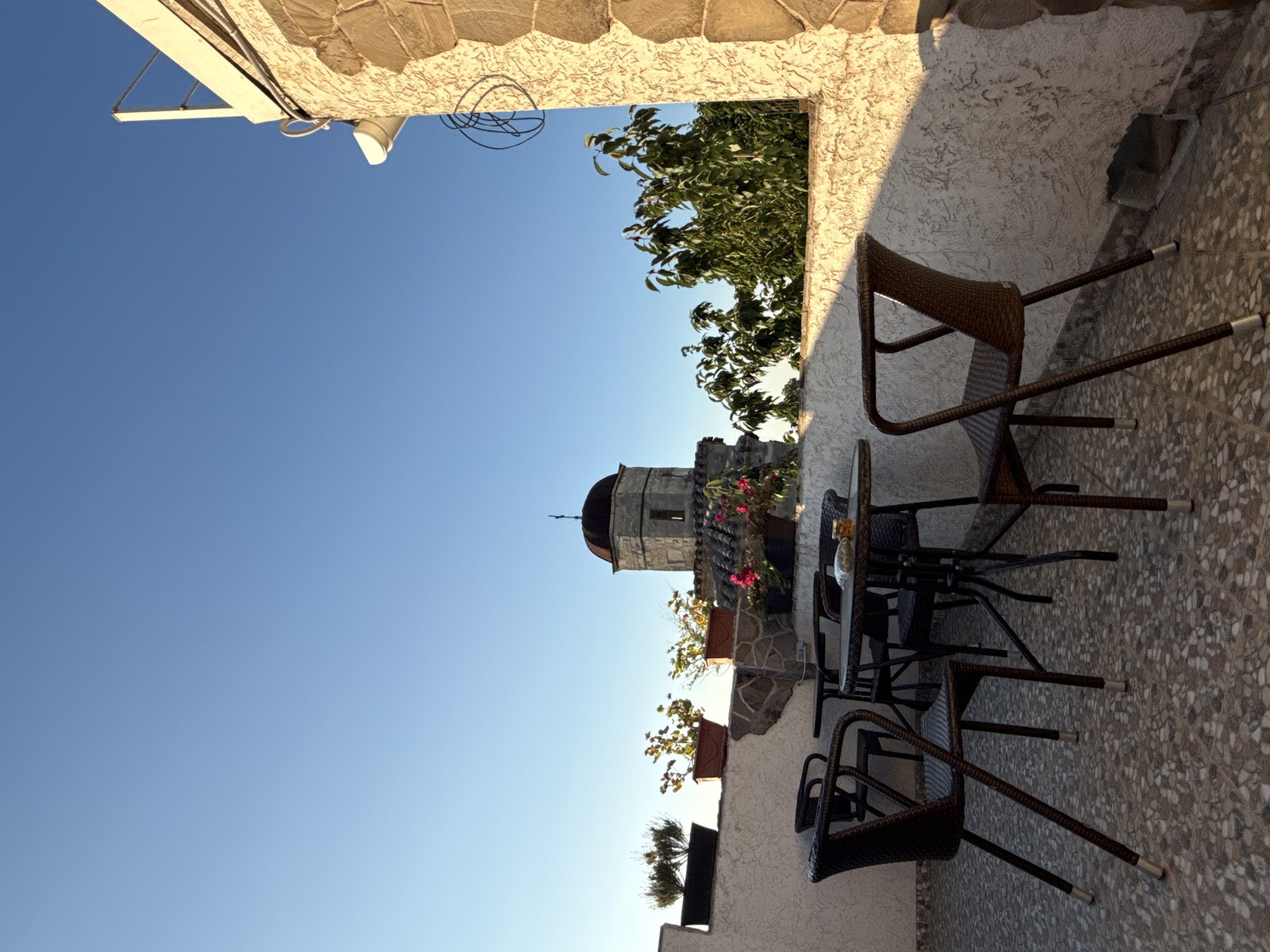 The image depicts an outdoor patio area in Feodosia, Ukraine, bathed in the warm light of what appears to be late afternoon or early morning sun. The sky is a clear, vibrant blue, dominating the upper portion of the frame. In the middle ground, a distinctive stone tower with a domed roof rises above other structures, its architecture suggesting a historical or religious building. Surrounding the tower are lush green trees and potted plants, some of which display pink flowers.
The foreground focuses on a small outdoor seating arrangement. A round table with a dark, possibly wicker or metal, surface is surrounded by several matching chairs. One chair is positioned at the table, and another is angled slightly away. The table has a small container with what appears to be food or snacks on its surface. The chairs have a woven texture and slender legs with silver-capped feet. The patio floor is covered in speckled, mosaic-like tiles.
To the right of the seating area, a textured, pale-colored wall is visible, constructed with large, rough-hewn stones on its upper section. This wall casts a distinct shadow from the setting or rising sun. The overall impression is one of a tranquil, inviting space, likely part of a private residence or a small establishment, offering a view of the city's architecture and natural surroundings.