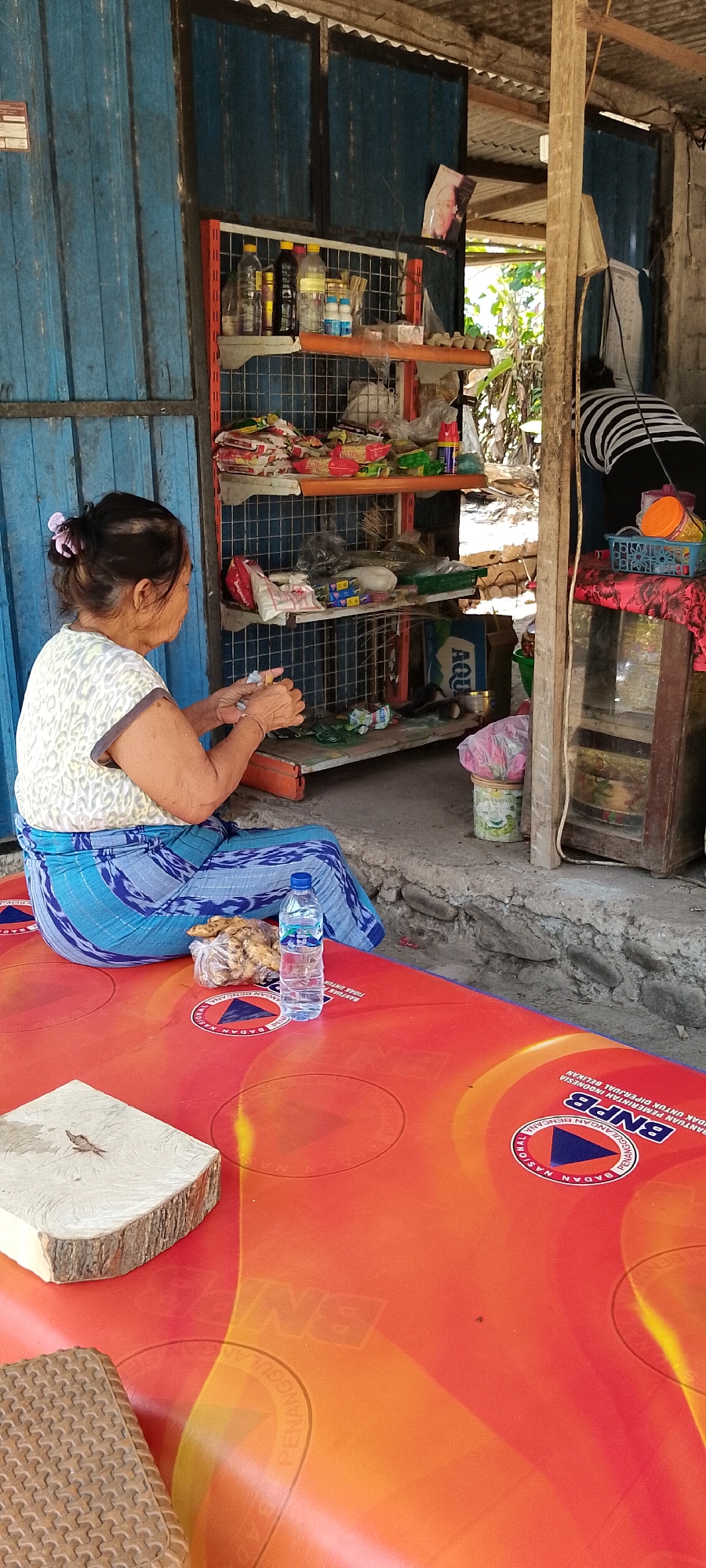 In Pekutatan, Indonesia, an elderly woman sits on a red mat in front of shelves stocked with various goods. She is wearing a white patterned t-shirt and a blue sarong. Her hands are occupied with what appears to be small items, possibly food or packaging. A plastic bottle of water and a bag of what looks like fried food are on the mat in front of her. Behind her, another person with black and white striped clothing is visible, facing away from the camera. The setting appears to be an outdoor stall or shop with blue wooden panels on the left and a corrugated metal roof. The shelves are filled with a variety of items, including bottles, packaged food, and what seem to be cartons. The ground is a mix of concrete and gravel. The red mat has circular patterns and text, including "BNPB," which is the Indonesian National Board for Disaster Management.