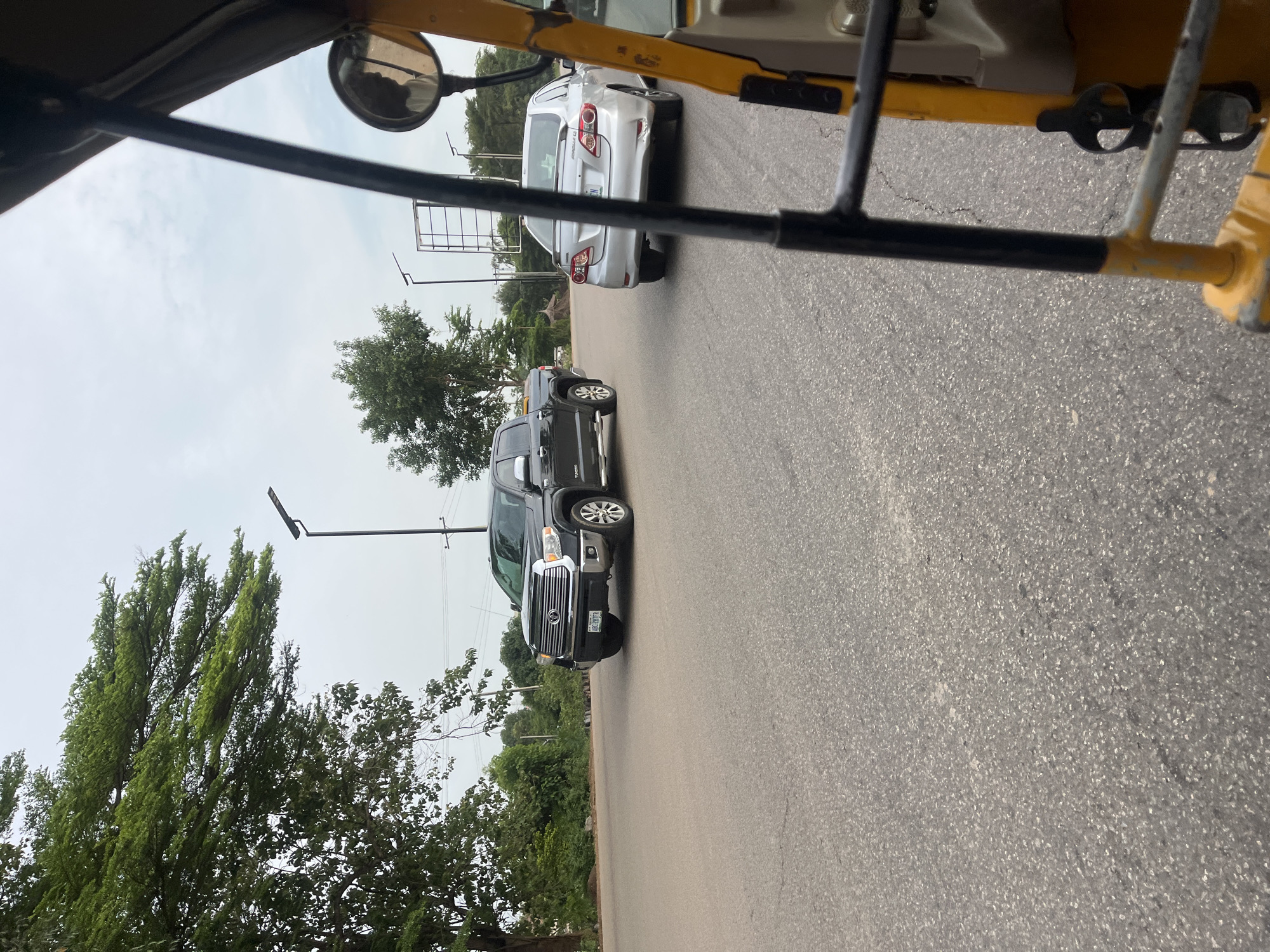 The image captures an outdoor street scene in Kuchingoro, Nigeria, taken from the interior of a vehicle, likely an auto-rickshaw, indicated by the visible yellow and black metal frame and a side-view mirror. The perspective looks out over a paved asphalt road beneath an overcast, bright sky.
In the midground, two vehicles are prominently featured. A silver Toyota Corolla sedan is seen from the rear, with the word "COROLLA" legible on its trunk. The rear left bumper of this car appears to be damaged or dented. Its license plate is partially visible, beginning with "ABJ 6..". Ahead of or beside the Corolla, a dark-colored pickup truck, identifiable by the "TOYOTA" badge on its front grille, is either parked or moving slowly. Its license plate, "ABJ 428WFA", is clearly visible.
The background features lush green trees lining the roadside, along with several utility poles or streetlights equipped with solar panels. The sky is uniformly bright but grey, suggesting an overcast daytime. The "ABJ" prefix on the vehicle license plates indicates the location is within Abuja, which is consistent with Kuchingoro being a district of the city. The scene depicts a common street environment with vehicles, but no people or specific activities are discernible.