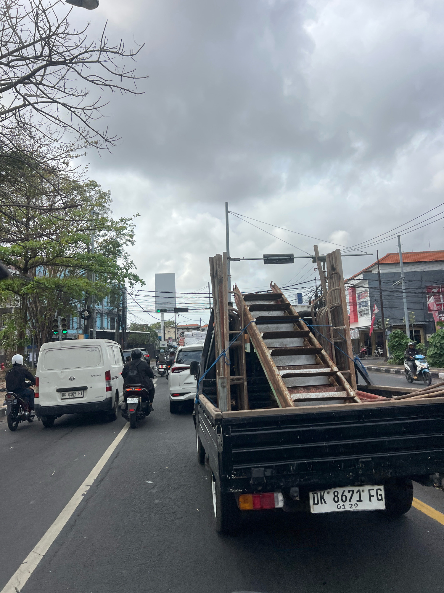 The image shows a street scene in Denpasar, Indonesia, with a black pickup truck carrying a metal staircase in its bed. The truck has a license plate that reads "DK 8671 FG". Several motorcycles are visible on the road, with riders wearing helmets. A white van with license plate "DK 8105 FI" is also present, along with other vehicles and buildings in the background. The sky is overcast with heavy clouds. The overall scene depicts traffic on a city street.