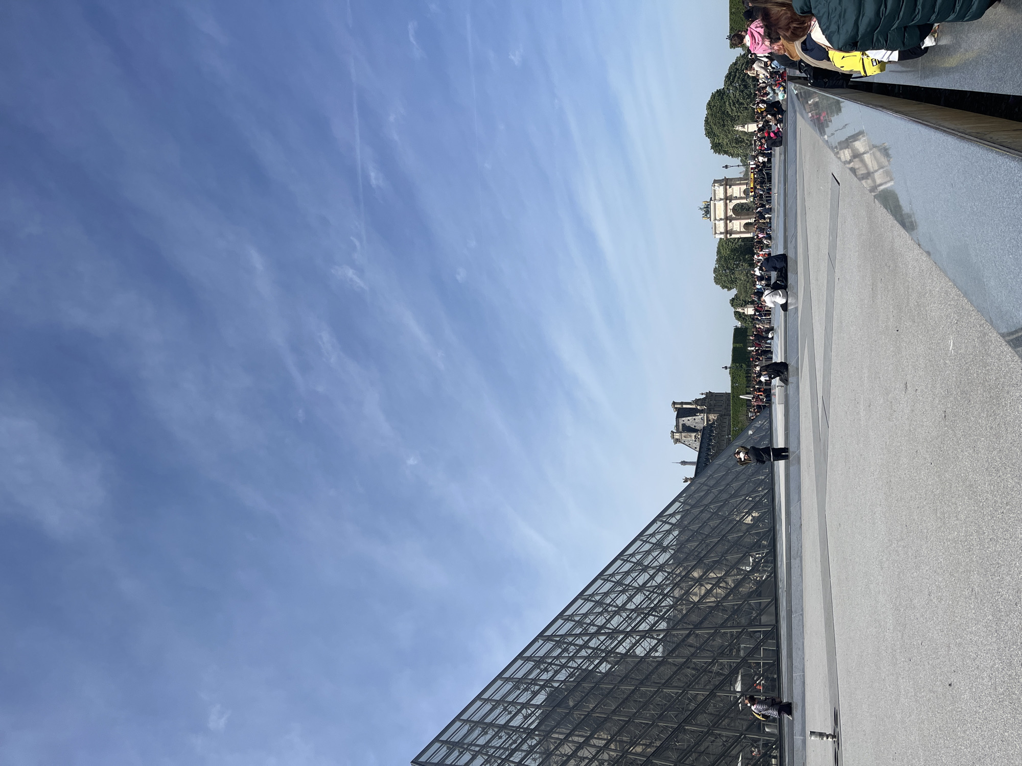 The image captures an outdoor scene at the Louvre Museum complex in Paris, France, on a bright, clear day. The iconic glass and metal Louvre Pyramid is prominently featured in the foreground, its intricate triangular structure reflecting the daylight. To the right of the pyramid, a wide, light gray paved area extends, with a walkway where numerous people are visible.
A considerable crowd of people, appearing to be tourists or visitors, are gathered and walking in the midground and background. They are distributed along pathways, some appearing to be in queues or congregating near the entrance to the museum. In the far background, behind the crowds and some trees, the Arc de Triomphe du Carrousel is clearly visible, showcasing its classical architecture. Further right, parts of the historic Louvre Palace buildings with their distinctive rooftops can be discerned.
The upper and left portions of the image are dominated by a vast, bright blue sky, with only a few thin, wispy white clouds scattered across it, indicating pleasant weather. The overall activity suggests a busy day at a popular tourist attraction. No visible text is present in the image.
