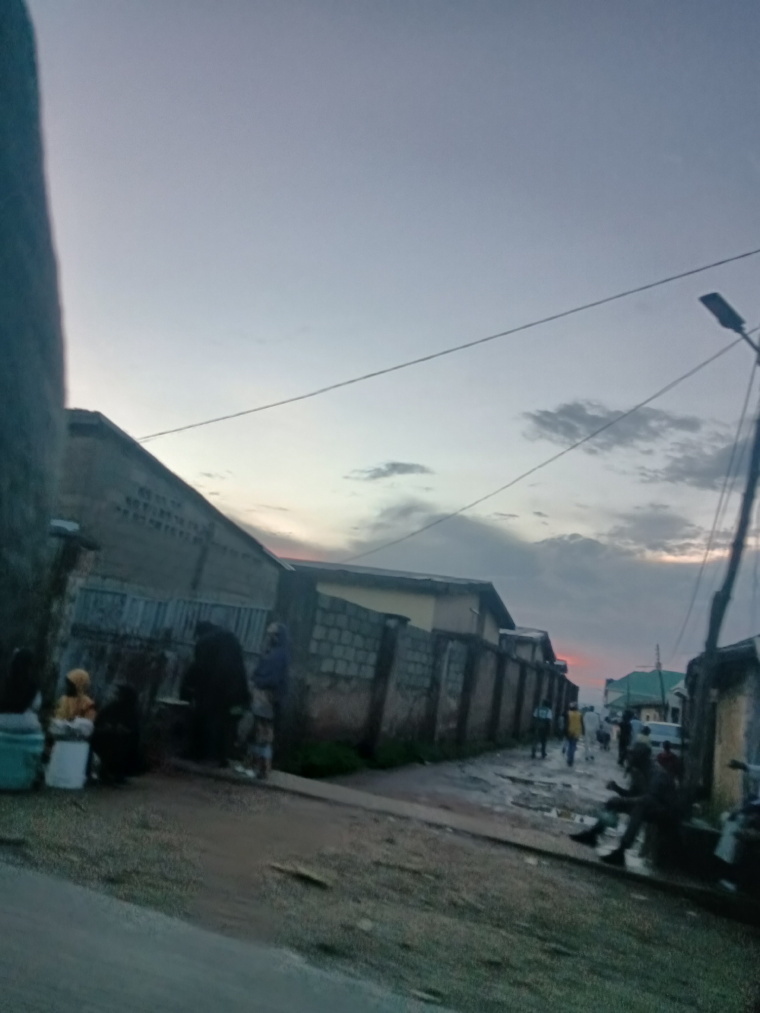 This image depicts a street scene in Jos, Nigeria at dusk. The sky is overcast with a gradient of pale blue and gray, with hints of orange and pink near the horizon. A street light stands tall on the right side of the frame, with wires strung across the sky. The street itself appears to be unpaved and somewhat wet, suggesting recent rain.
Buildings line both sides of the street. On the left, a low-rise building with a corrugated metal roof and brick walls is visible. On the right, another building with a yellow facade and a green roof is partly visible. The area seems to be a residential or mixed-use district, with some areas appearing to be under construction or in disrepair, indicated by exposed brickwork and uneven ground.
Several people are present in the scene. In the foreground on the left, a group of individuals are gathered, some sitting or crouching near what appear to be containers. Further down the street, other individuals are walking or standing, some in small groups. The activity suggests daily life in the neighborhood, possibly involving street vending or informal gatherings. The overall atmosphere is muted, characteristic of the twilight hours in a busy urban environment.