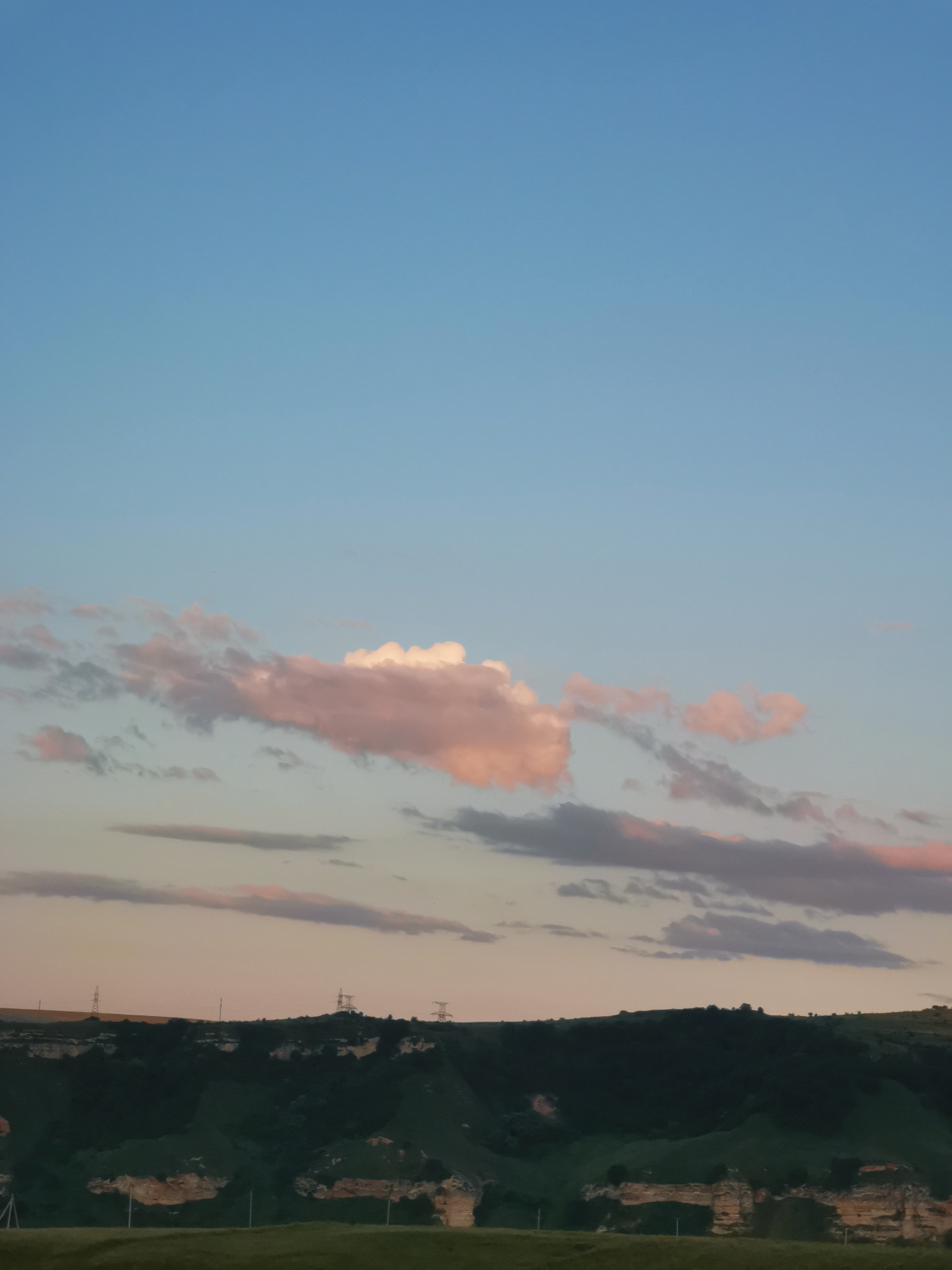 The image depicts a serene landscape under a pale blue sky with scattered clouds tinged with pink and orange hues, suggesting either sunrise or sunset. The horizon is defined by a dark, undulating silhouette of hills or mountains, sparsely dotted with what appear to be trees or shrubs. Some of these hills exhibit exposed rock formations with a sandy or light brown color. Electrical pylons are visible along the ridge of the hills, indicating the presence of human infrastructure. The foreground shows a gently sloping green field, with hints of other faint structures or markers, possibly fences. The overall scene is tranquil and natural, with a sense of open space. The location is identified as Kubina, Russia.