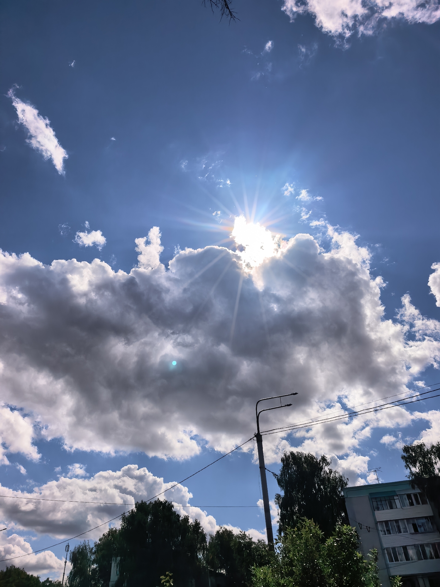 The image depicts a bright, sunny day with a vibrant blue sky filled with scattered white and grey clouds. The sun is positioned high in the sky, partially obscured by a large, dense cloud, casting brilliant rays and a lens flare. In the lower portion of the image, a street lamp stands tall, with power lines stretching across the frame. Beyond the lamp and wires, lush green trees are visible, and to the right, a multi-story apartment building with visible windows can be seen. This scene is located in the city of Sterlitamak, Russia, suggesting an urban or suburban environment. There are no people or animals visible in the frame.