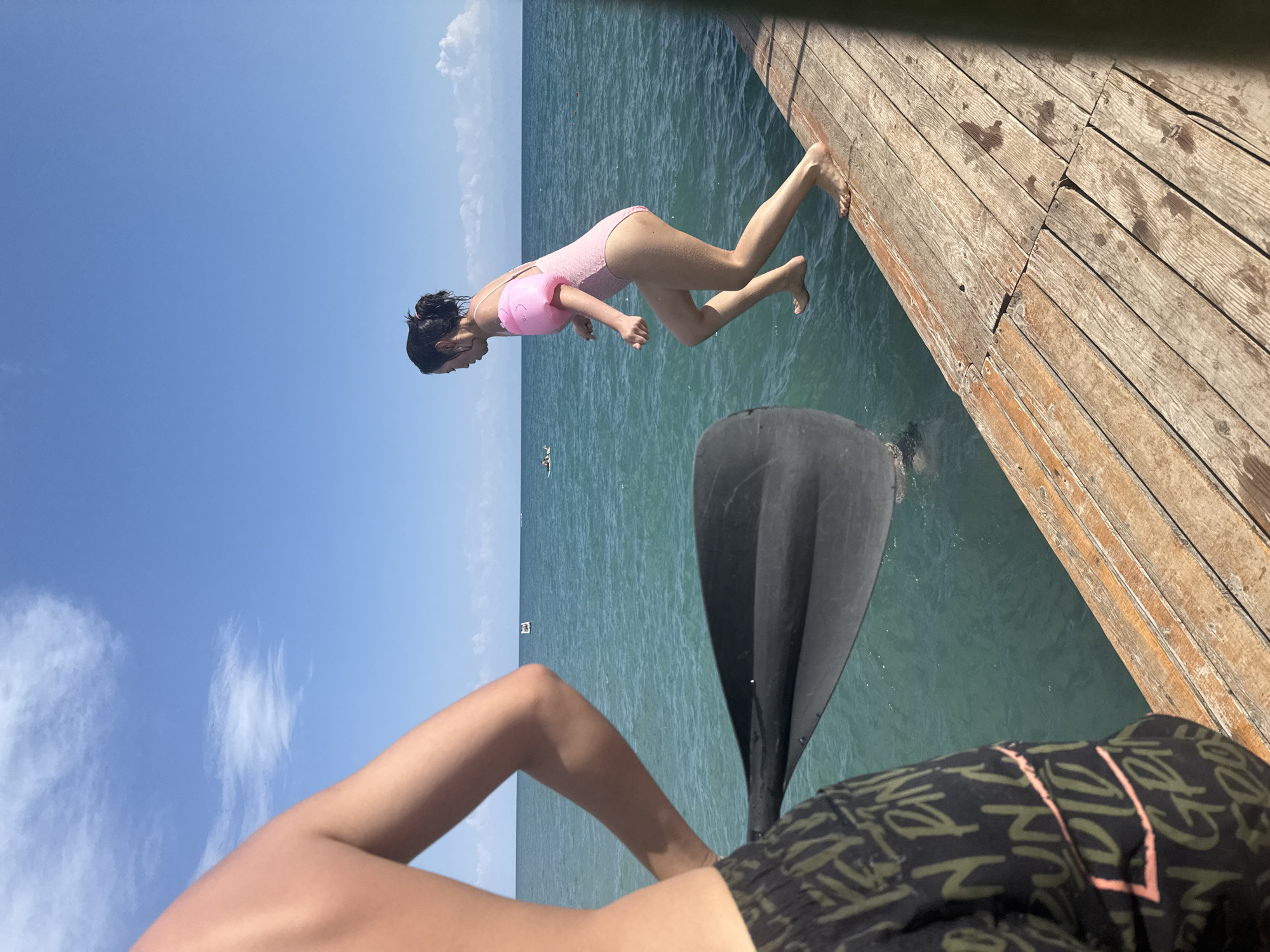 In the city of Sary-Oy, Kyrgyzstan, a young woman wearing a pink one-piece swimsuit with inflatable armbands is captured mid-jump from a wooden pier into the turquoise waters of Issyk-Kul Lake. She is poised with her body in motion, right leg bent and left leg extended, preparing to enter the water. Her hair is tied back in a bun. In the foreground, the back of another person is visible, partially obscuring the view with their arm and a black paddle. This person is wearing black swim shorts with a camouflage pattern and various green and pink text. The pier, constructed of weathered wooden planks, runs along the right side of the image. The clear blue sky, dotted with white clouds, stretches across the horizon, meeting the calm lake. In the distance, a few small boats are visible on the water.