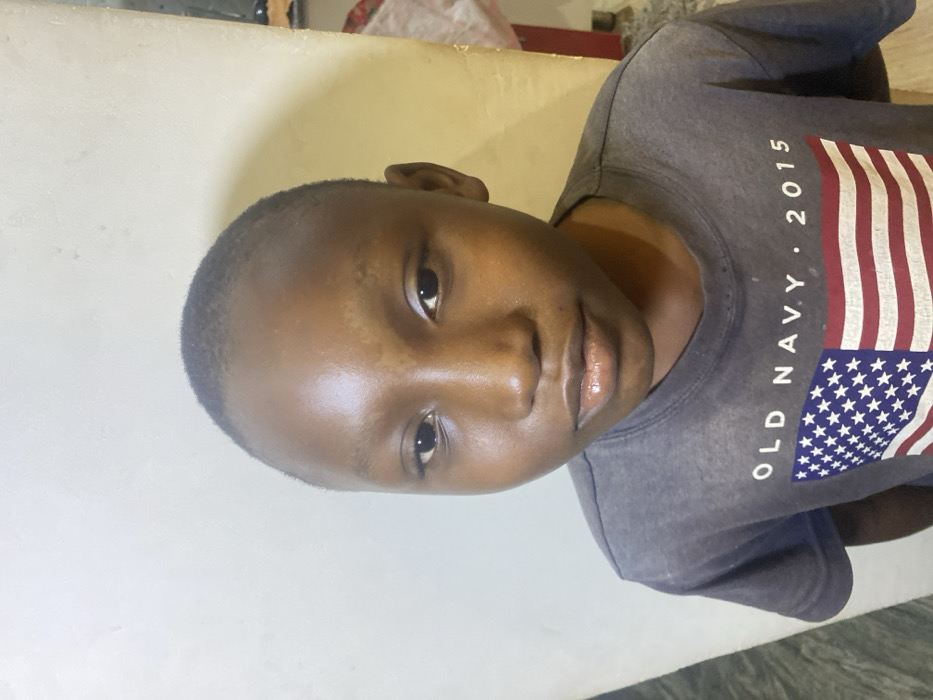 A close-up, slightly tilted shot features a young boy with short, dark hair. He has smooth, dark skin and dark eyes, looking directly at the camera with a gentle expression. He's wearing a gray t-shirt featuring a stylized American flag design along with white lettering that reads "OLD NAVY. 2015". The background consists of a plain, light-colored wall.