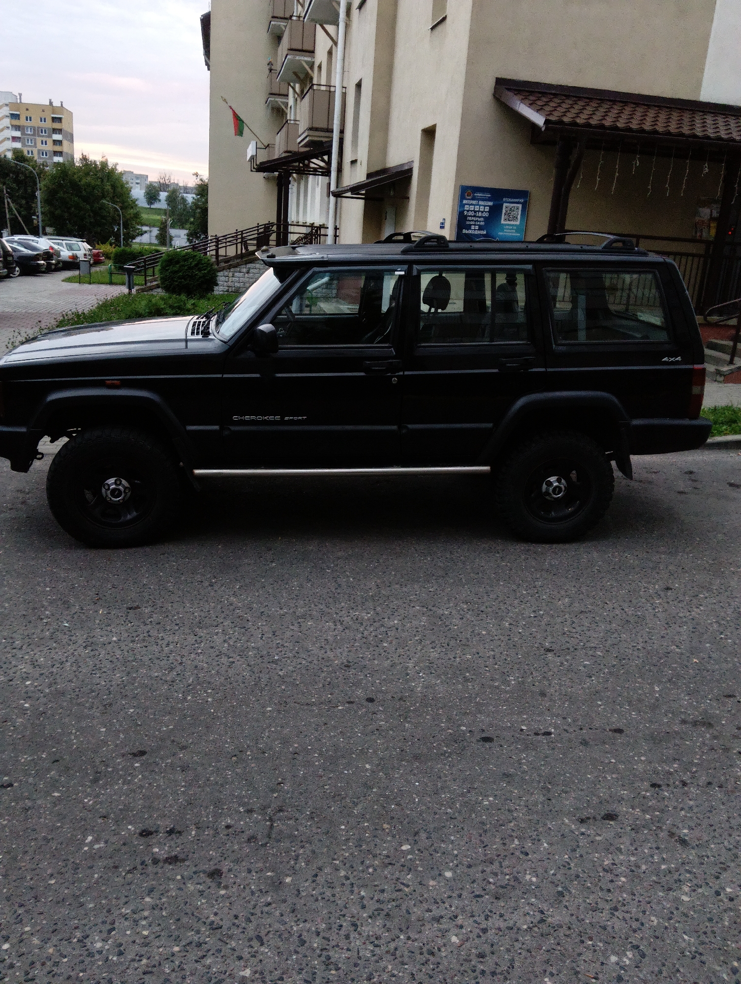 A black Jeep Cherokee Sport is parked on a paved asphalt road in front of a multi-story building. The building is light beige with balconies and a dark tiled roof. A sign with blue lettering and a QR code is visible on the building's facade. A Belarusian flag, red and green, flies on a pole attached to the building. To the left of the Jeep, other cars are parked on a paved area. Green trees and distant buildings are visible in the background under an overcast sky. The scene appears to be in a residential area of Hrodna, Belarus.