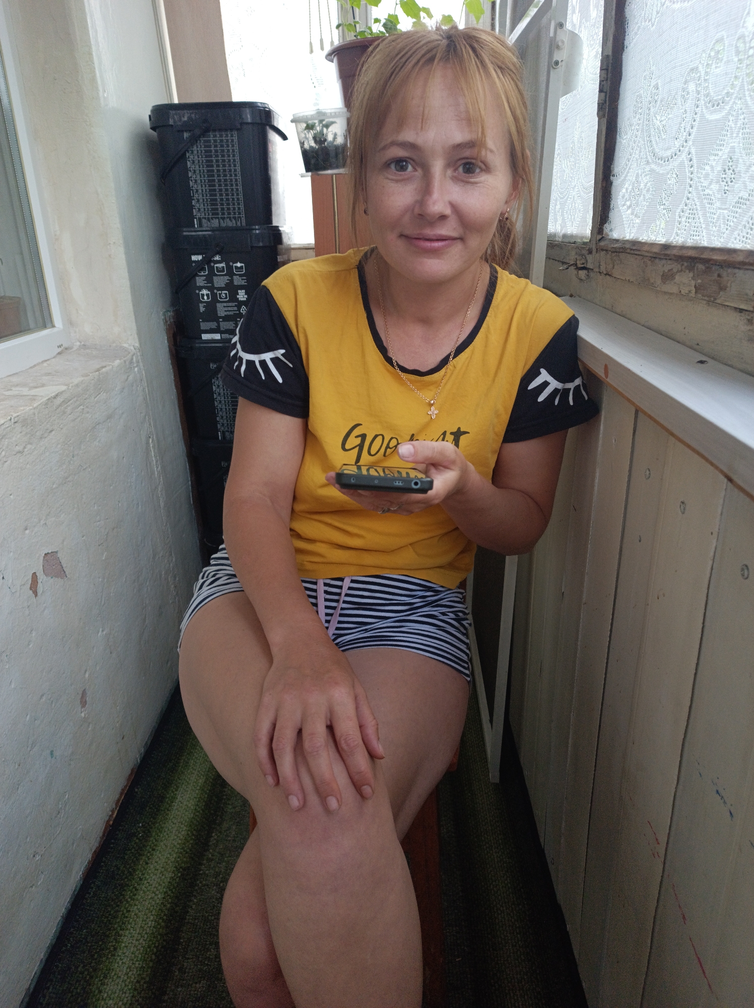 A woman with blonde hair is sitting on a stool, holding a mobile phone in her right hand. She is wearing a yellow t-shirt with black sleeves and black and white striped shorts. The t-shirt has a graphic of an eye with eyelashes on each sleeve and text on the front that is partially visible. She is wearing a gold necklace with a cross pendant.
The background shows a white wall on the left, and a wooden wall on the right. There are some black buckets stacked up behind the woman. A window with a white lace curtain is visible behind the woman and to her right. There are some plants in pots on a windowsill above the wooden wall. The scene appears to be indoors, on a balcony or enclosed porch. The lighting suggests it is daytime. The woman's expression is neutral.
The text on the t-shirt is difficult to read, but the word "GOOD" is visible.