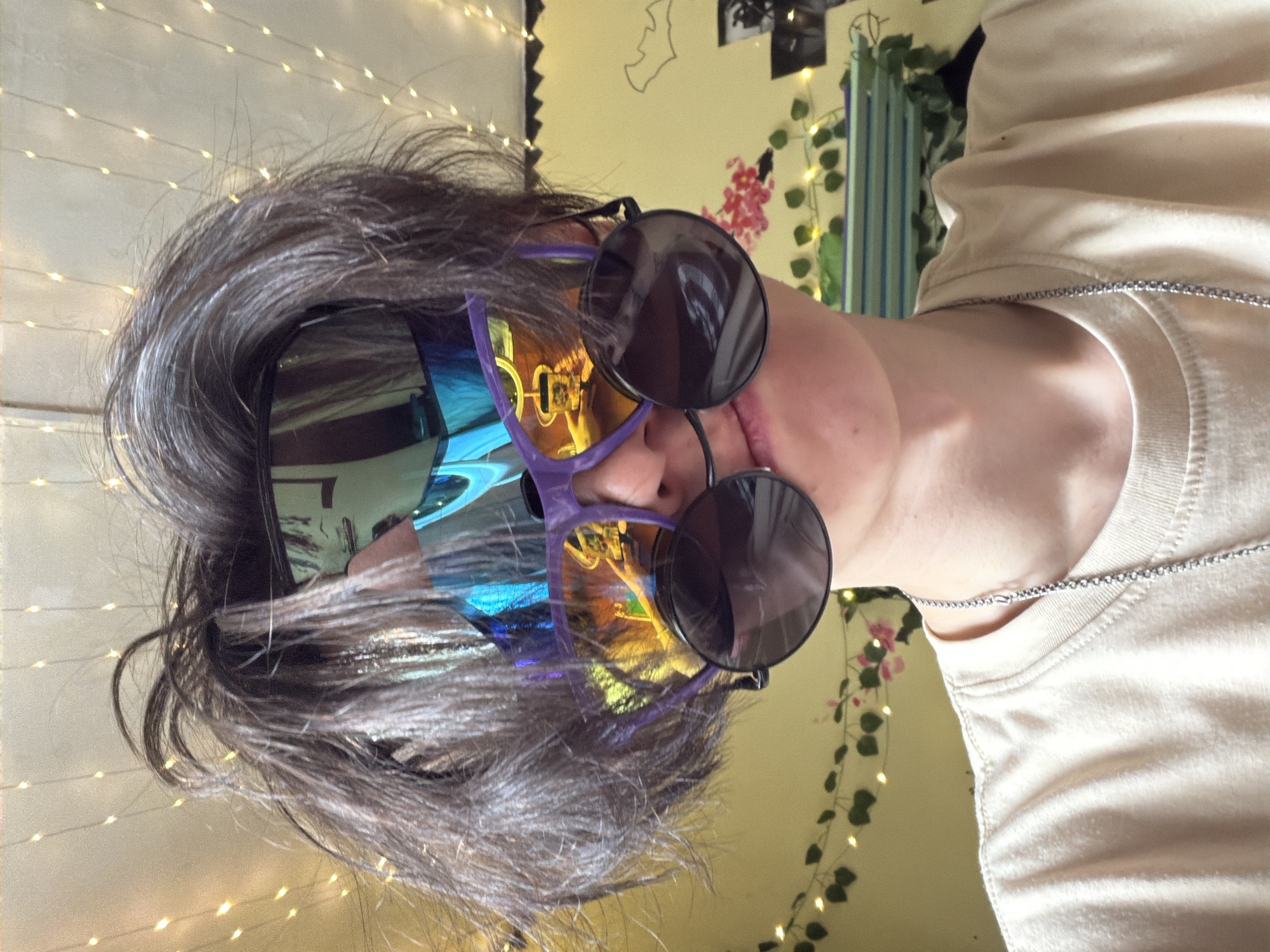 The media file is a selfie taken from a low angle, showing a person wearing large, colorful sunglasses. The person has dark, messy hair that falls over their face, partially obscuring their features. The sunglasses have purple frames and reflective lenses that show a distorted image of the surroundings, including what appears to be a room with fairy lights and some wall decorations. The person is wearing a light-colored t-shirt with a thin chain necklace. The background is a wall decorated with string lights, artificial greenery, and some floral or abstract art. The lighting is dim, suggesting it might be nighttime or an indoor setting. The overall mood is playful and casual, with the exaggerated sunglasses adding a quirky element.