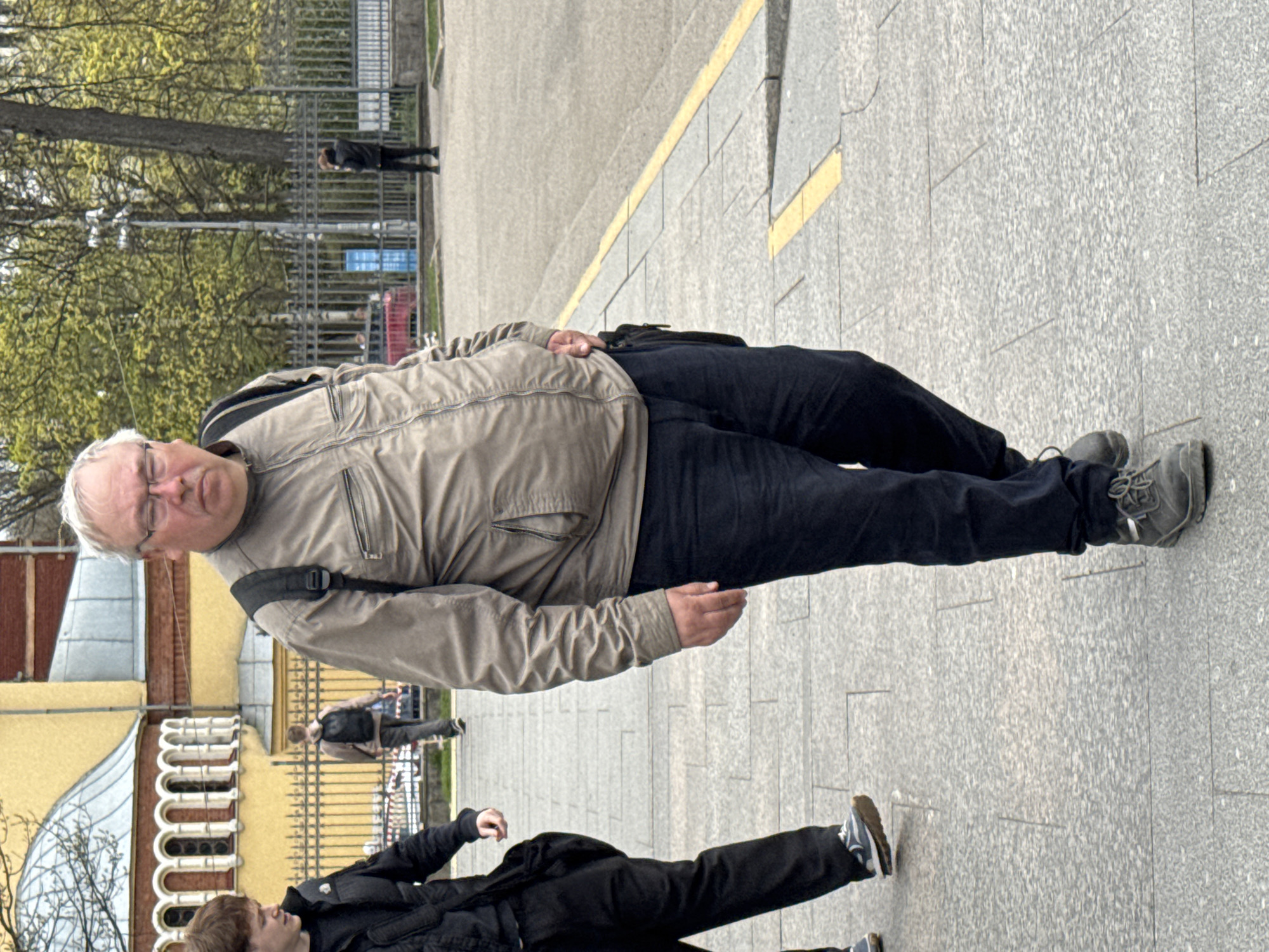 An eye-level shot shows a medium-built middle-aged white man with graying hair and glasses, wearing a brown jacket and dark pants and shoes, standing on a light gray tiled sidewalk. A yellow line runs along the top of the sidewalk. A person wearing a black jacket and pants is standing and pointing at the bottom of the photo. Other pedestrians are visible walking behind a light yellow wrought iron fence with arched openings, next to a building with a half-moon-shaped metal-covered roof. The location appears to be Saint Petersburg, Russia, based on the architecture and streetscape. A tree and metal fence are visible in the background.