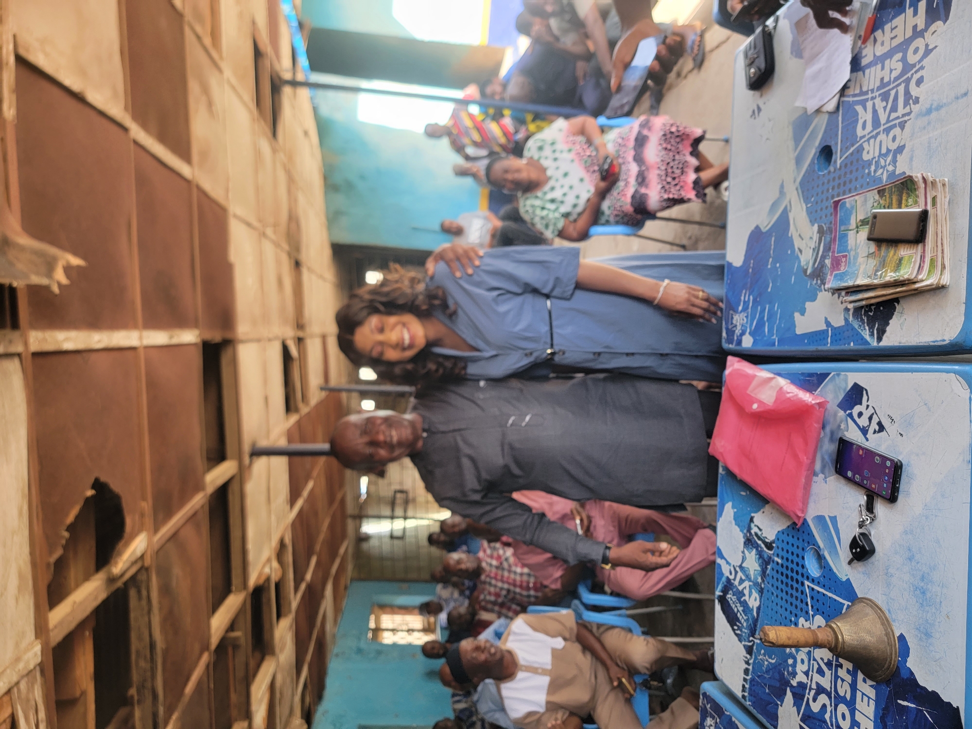 The image depicts a gathering in what appears to be a community hall in Sardauna, Nigeria. The hall has blue walls and a rusty brown metal and wooden structure. A group of people are seated in blue plastic chairs. A man and a woman are standing close together near the front of the room. The woman is smiling and has her arm around the man. A table is set up with items including a mobile phone, keys, a pink envelope, a pile of documents, and a brass bell.