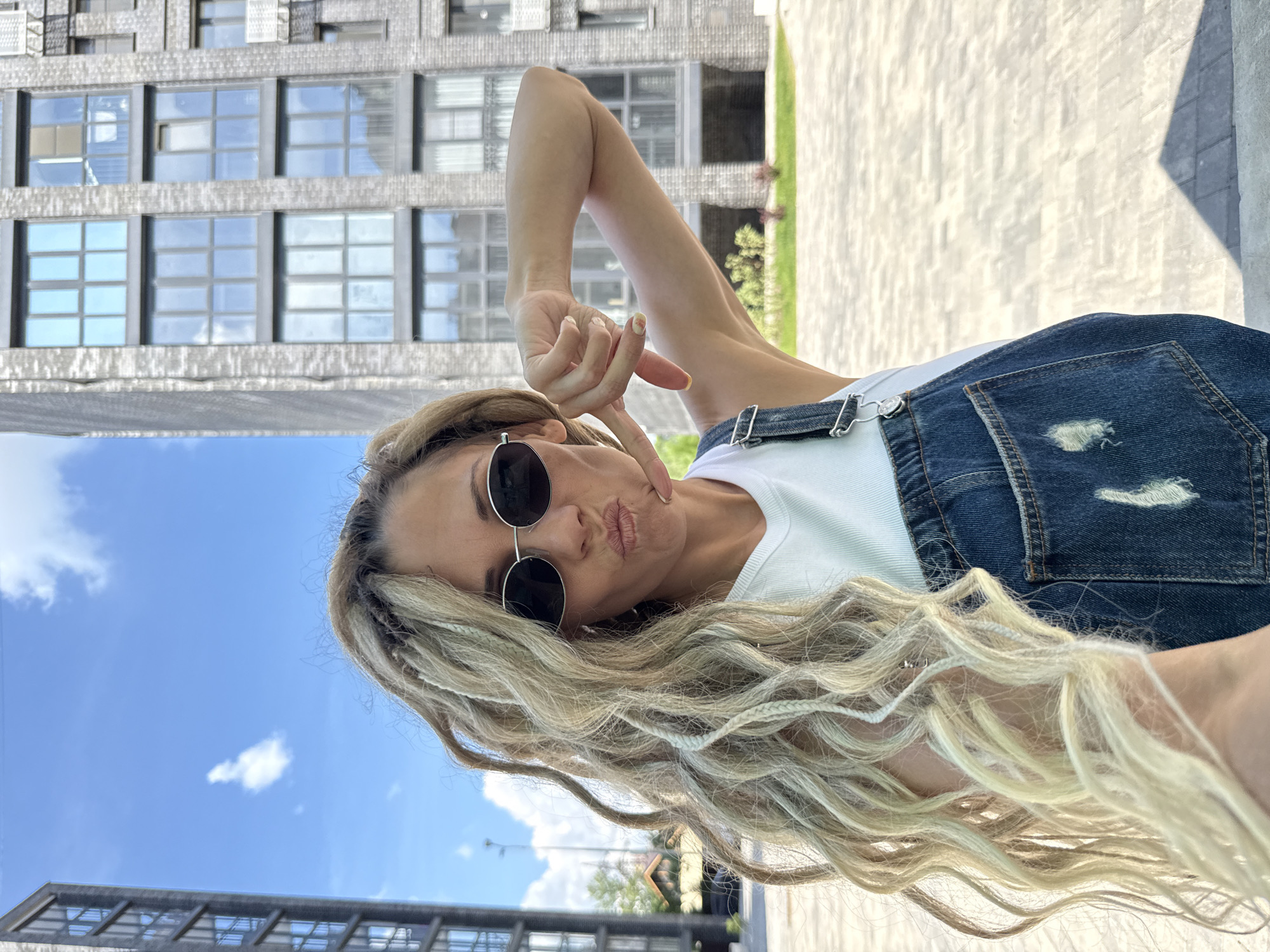 A woman with blonde dreadlocks and sunglasses is taking a selfie outdoors in front of a modern brick building with large windows. She is wearing a white top and denim overalls. She is making a kissing face and gesturing with her hand. The background shows a clear blue sky with some clouds, the building, and a paved area. The location is identified as Rusanovo, Russia.