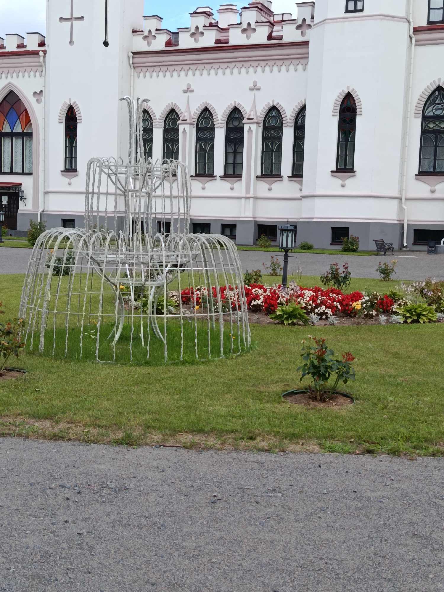 The image shows the Kosava Castle in Kosava, Belarus. The castle is a large, white building with a distinctive architectural style, featuring pointed arches, crenellations, and stained-glass windows. In front of the castle, there is a manicured lawn with a decorative metal structure that resembles a fountain. A flower bed filled with red and white flowers is also visible, along with a lamppost and a park bench. The scene appears to be set on a cloudy day, with the sky visible in the background. The image is captured from a ground-level perspective, looking towards the castle.