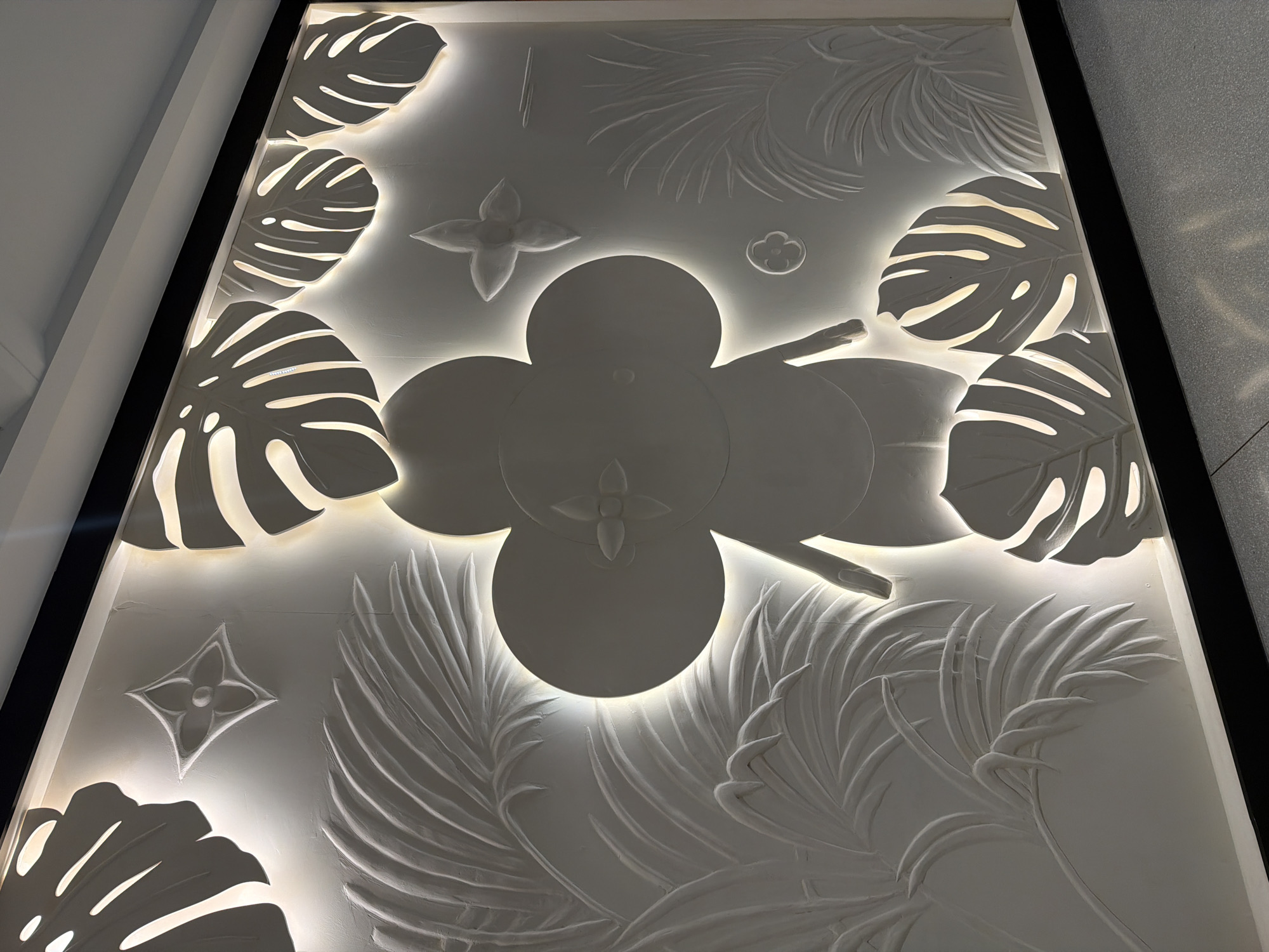 The image shows a close-up of a large, illuminated relief sculpture mounted on a wall. The sculpture features stylized botanical elements, including large Monstera leaves and palm fronds, rendered in white. A prominent central motif appears to be a stylized cartoon character's head and torso, with large, rounded ears and a flower-shaped emblem on its chest. Smaller floral emblems are also scattered throughout the composition. The lighting emanates from behind the sculpture, creating a dramatic halo effect around the raised elements and casting shadows that accentuate the dimensionality of the relief. The overall aesthetic suggests a modern, luxury brand installation. This artwork is located in the Louis Vuitton boutique in ICONSIAM, a luxury shopping mall on the banks of the Chao Phraya River in Bangkok, Thailand.