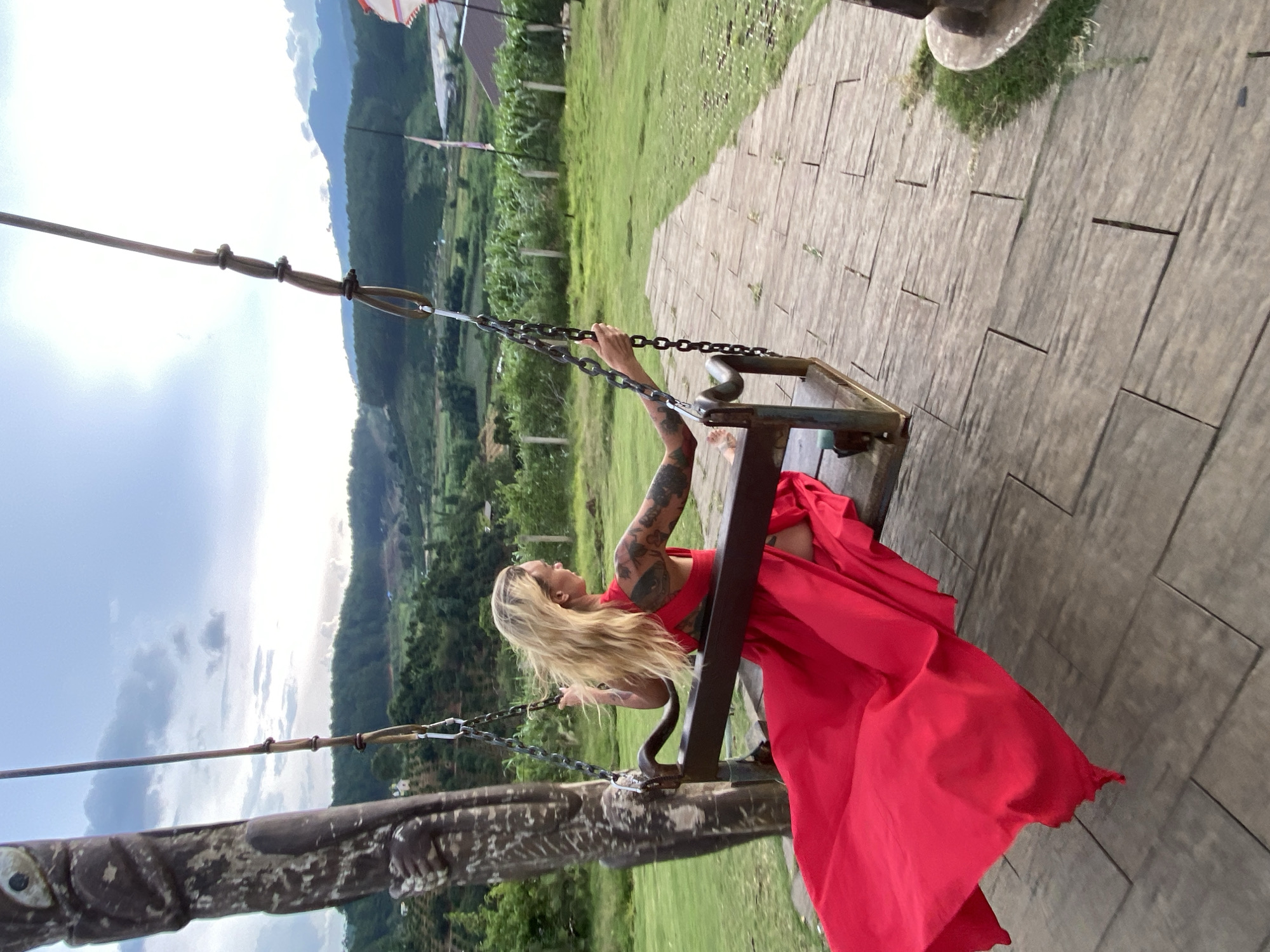 A woman with long blonde hair and tattoos on her arms is sitting on a swing. She is wearing a long, flowing red dress. The swing is made of metal and wood, and it is suspended by chains. The woman is holding onto the chains with her hands. She is looking off to the side, and her expression is serene.
The background of the image is a lush green landscape with rolling hills. There are some trees and a cloudy sky. The ground is covered in paving stones. The image appears to have been taken during the daytime. The weather is overcast.
The woman's red dress is vibrant and contrasts with the green landscape. The tattoos on her arms add an interesting detail to her appearance. The swing is rustic and complements the natural setting. The overall impression is one of peacefulness and natural beauty.