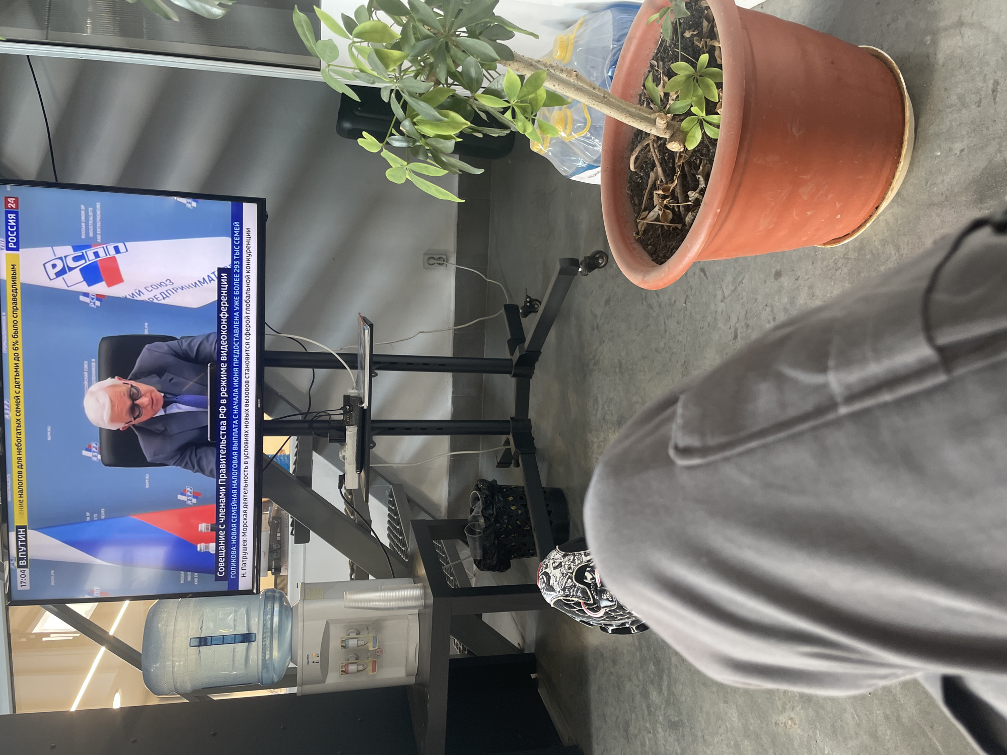 The image captures a casual moment in an office environment, with a television screen displaying a news broadcast from Russia 24. The broadcast features a man in a suit and glasses speaking, with the Russian flag and the logo of the Russian Union of Industrialists and Entrepreneurs visible in the background. Below the broadcast, a text overlay reads "Совещание с членами Правительства РФ в режиме видеоконференции" (Meeting with members of the Government of the Russian Federation in video conference mode). The foreground is dominated by the legs of a person wearing gray sweatpants and an athletic shoe. To the left of the television, a water cooler with a large bottle of water is situated on a black table. Nearby, a trash can and a potted plant with green leaves in a terracotta pot add to the office setting. The overall scene suggests a workspace in Sarapul, Russia, where an individual is taking a break to watch the news.