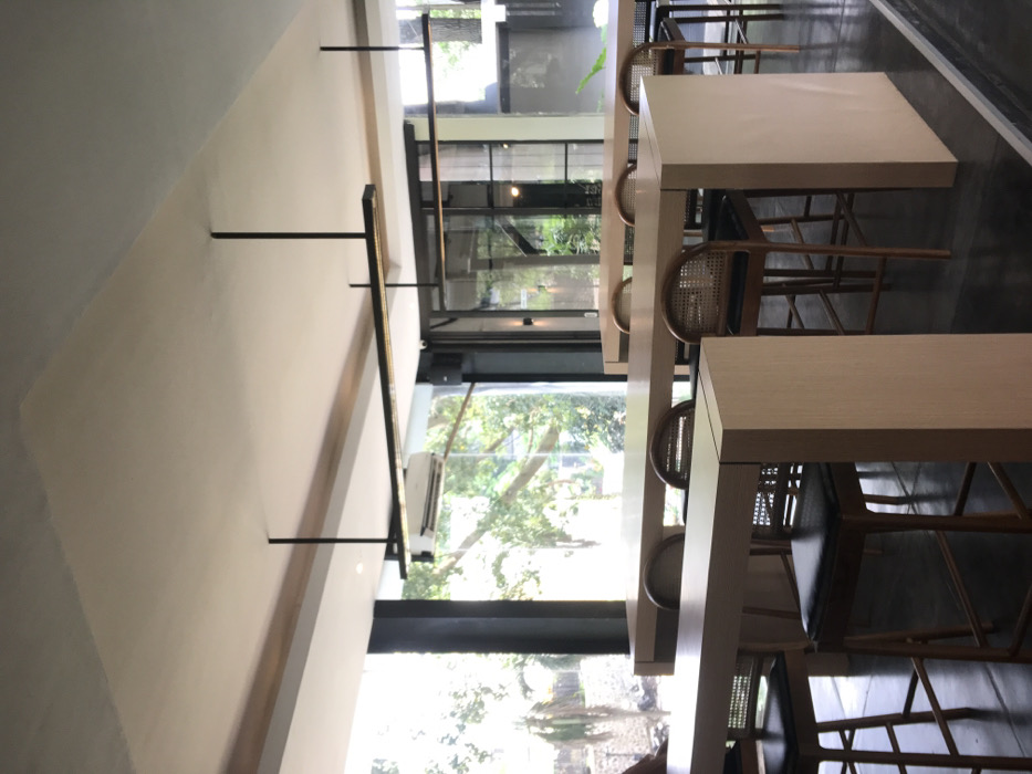 The image shows the interior of a modern restaurant located at coordinates -7.98 latitude and 112.617 longitude, placing it in Malang, Indonesia. The restaurant has a white ceiling with two long, black, horizontal light fixtures hanging from it. The floor is dark grey tile and the walls are painted white. Three long, light-brown wooden tables line the far wall. Each table is set with four black leather-backed chairs with woven cane seats. The tables are placed near a large window that looks out onto a green, leafy outdoor area. There are no people visible in the image. The space appears to be a restaurant or cafe. The tables and chairs suggest that it is a place for dining. The modern design and the bright lighting suggest that it is a contemporary establishment. The cleanliness and well-kept appearance suggest that it is well-maintained and popular.